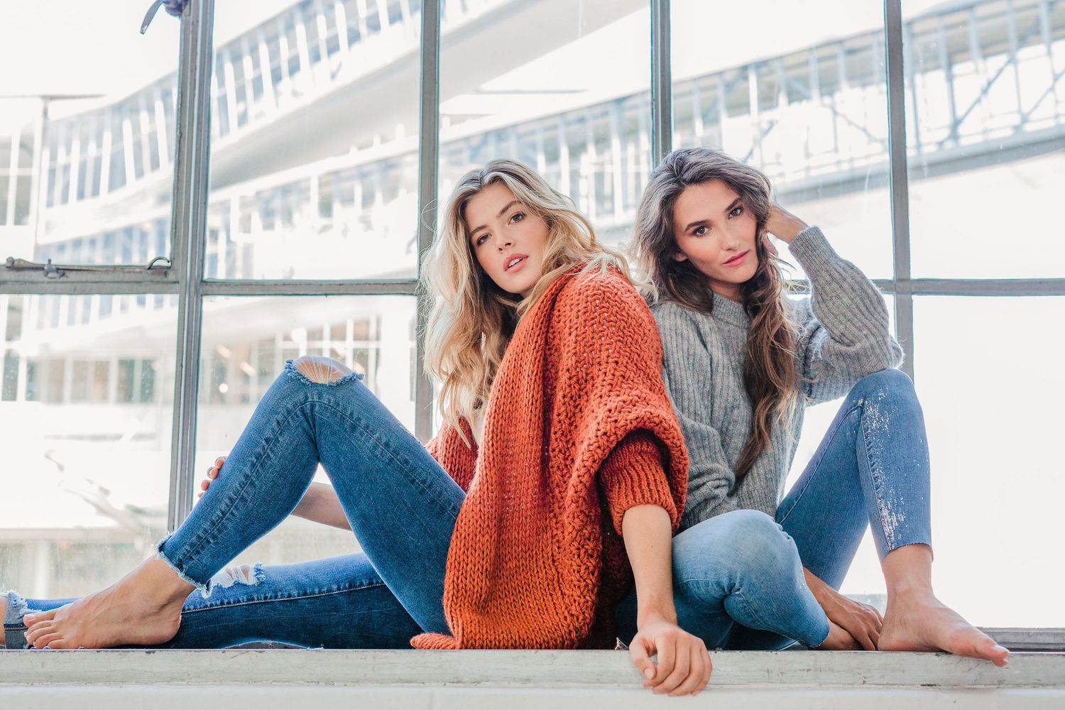 Couple in casual attire lounging by bright industrial windows in artistic photo series.