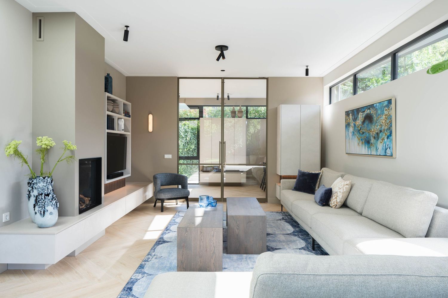 Modern living room with beige sectional sofa, blue accents, and floor-to-ceiling windows overlooking greenery.
