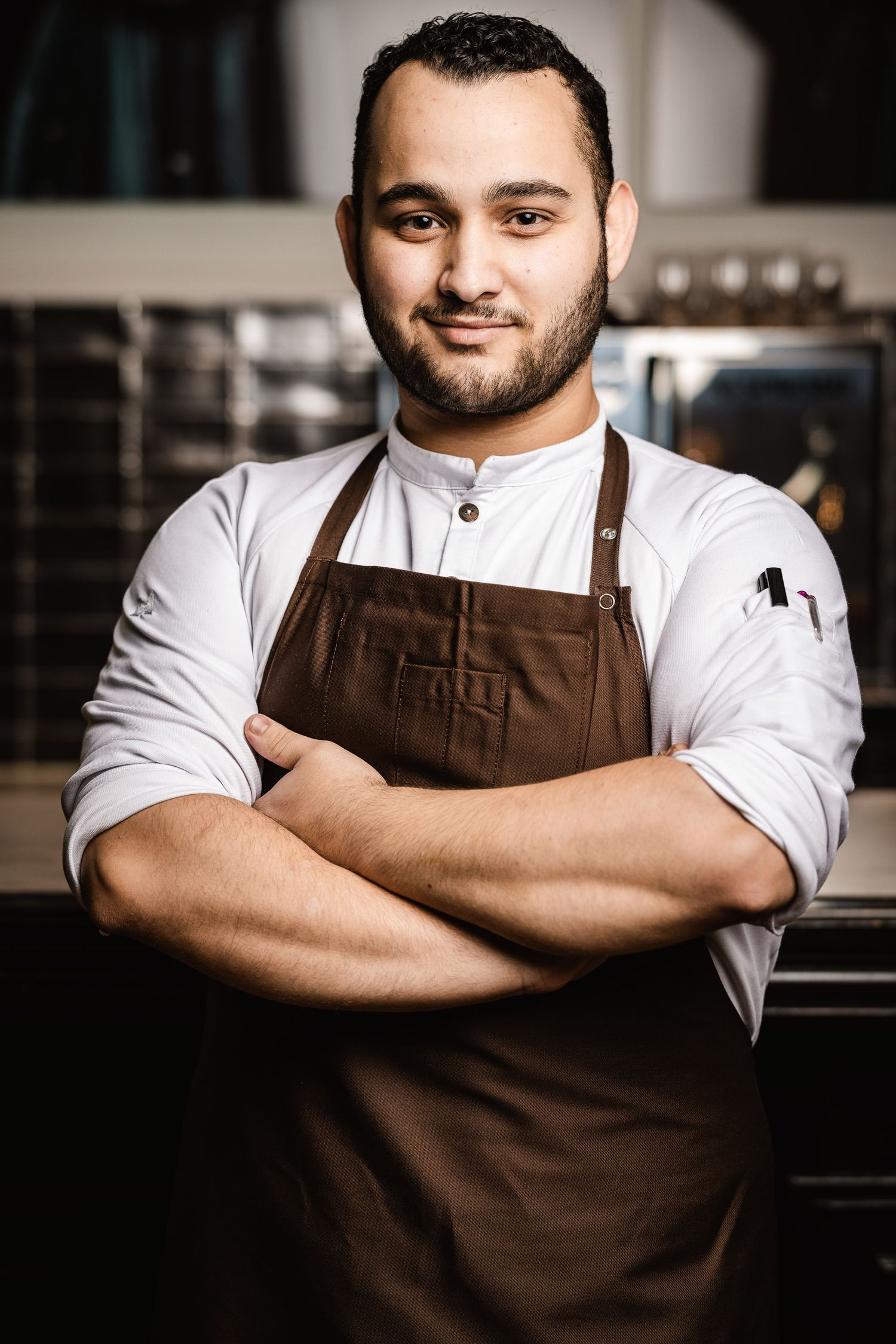 Chef in white shirt and brown apron poses with crossed arms in professional kitchen.