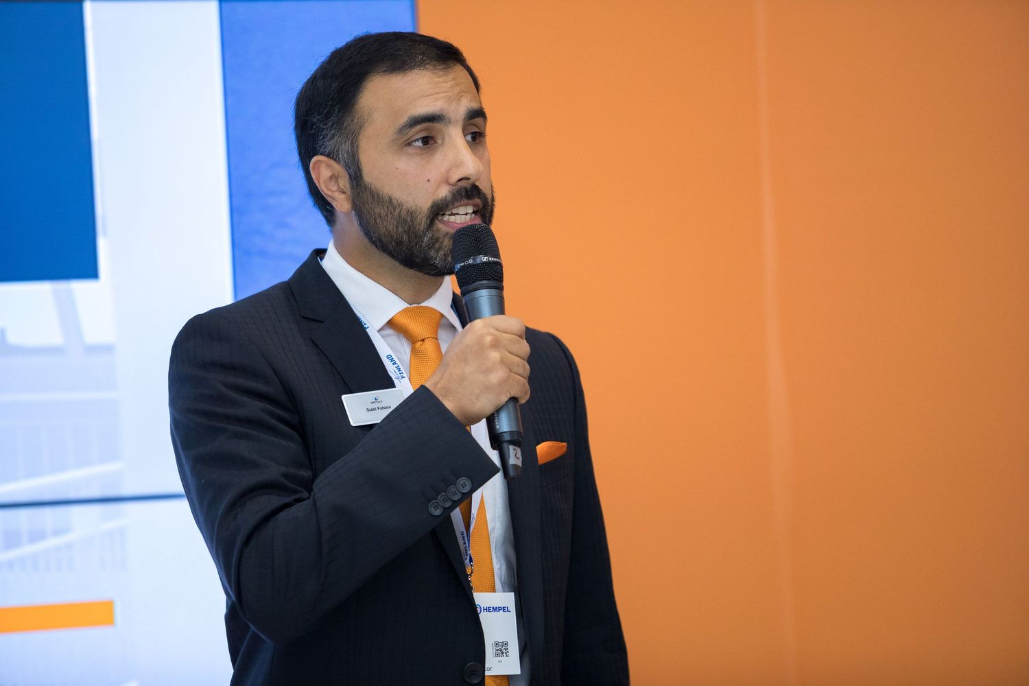 A speaker wearing a dark suit presents at a professional conference against an orange backdrop.