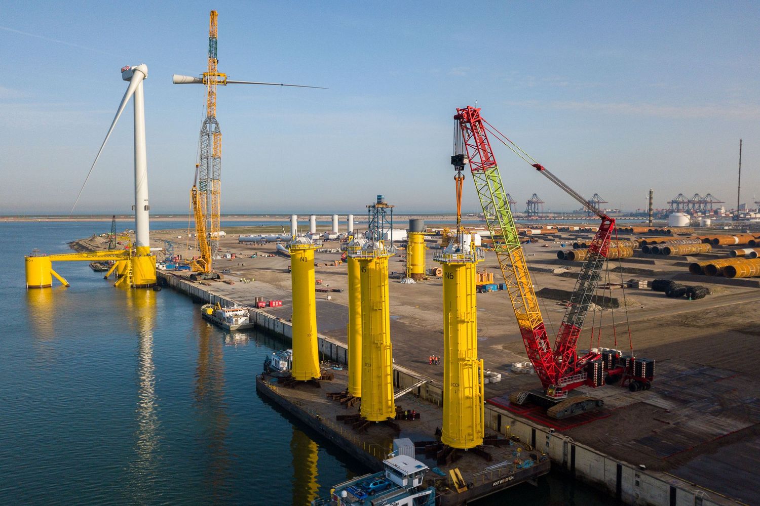 Industrial cranes and wind turbine components being assembled at a coastal port facility.