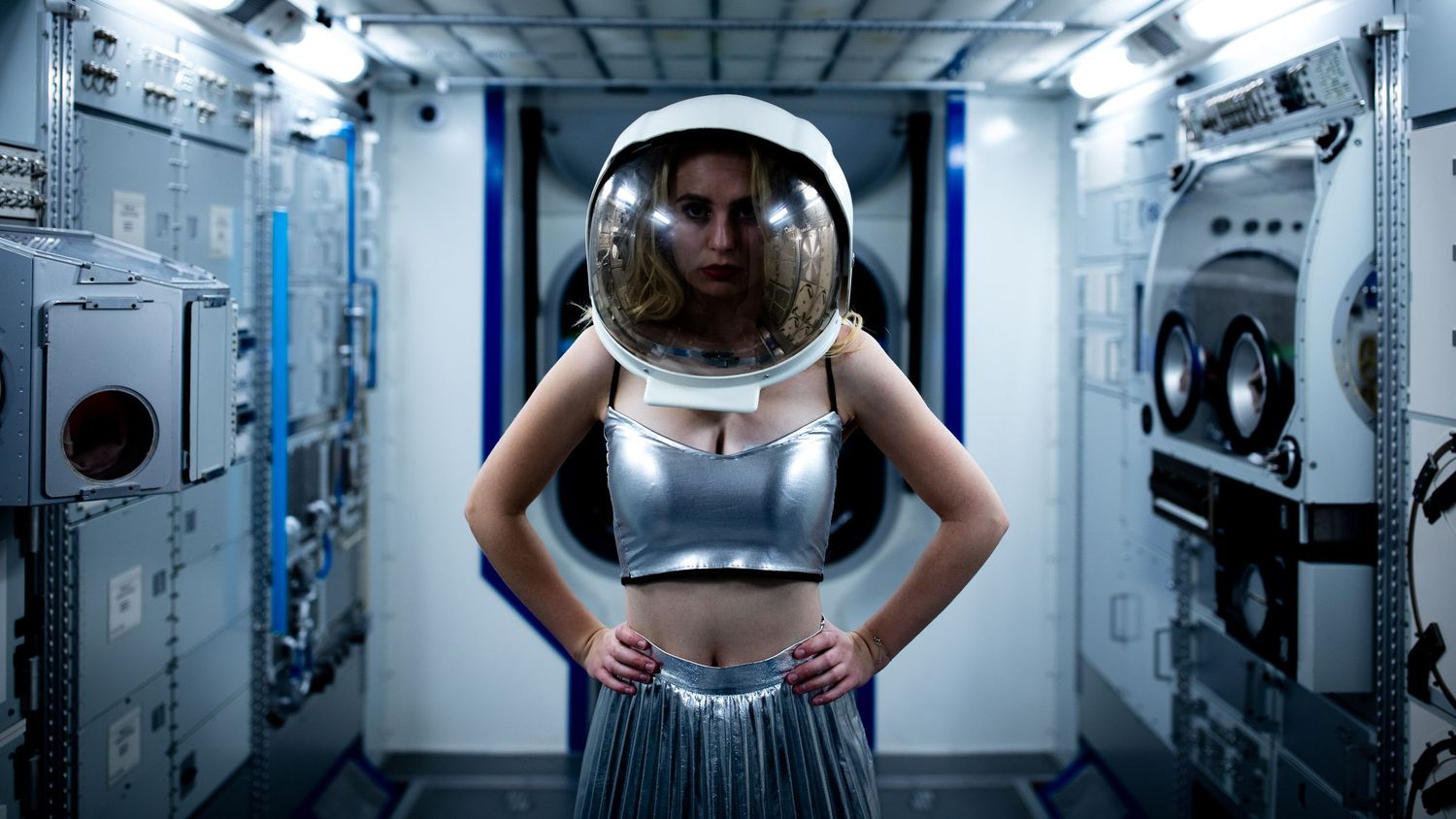 Woman in silver crop top and astronaut helmet poses confidently inside a futuristic space station corridor.