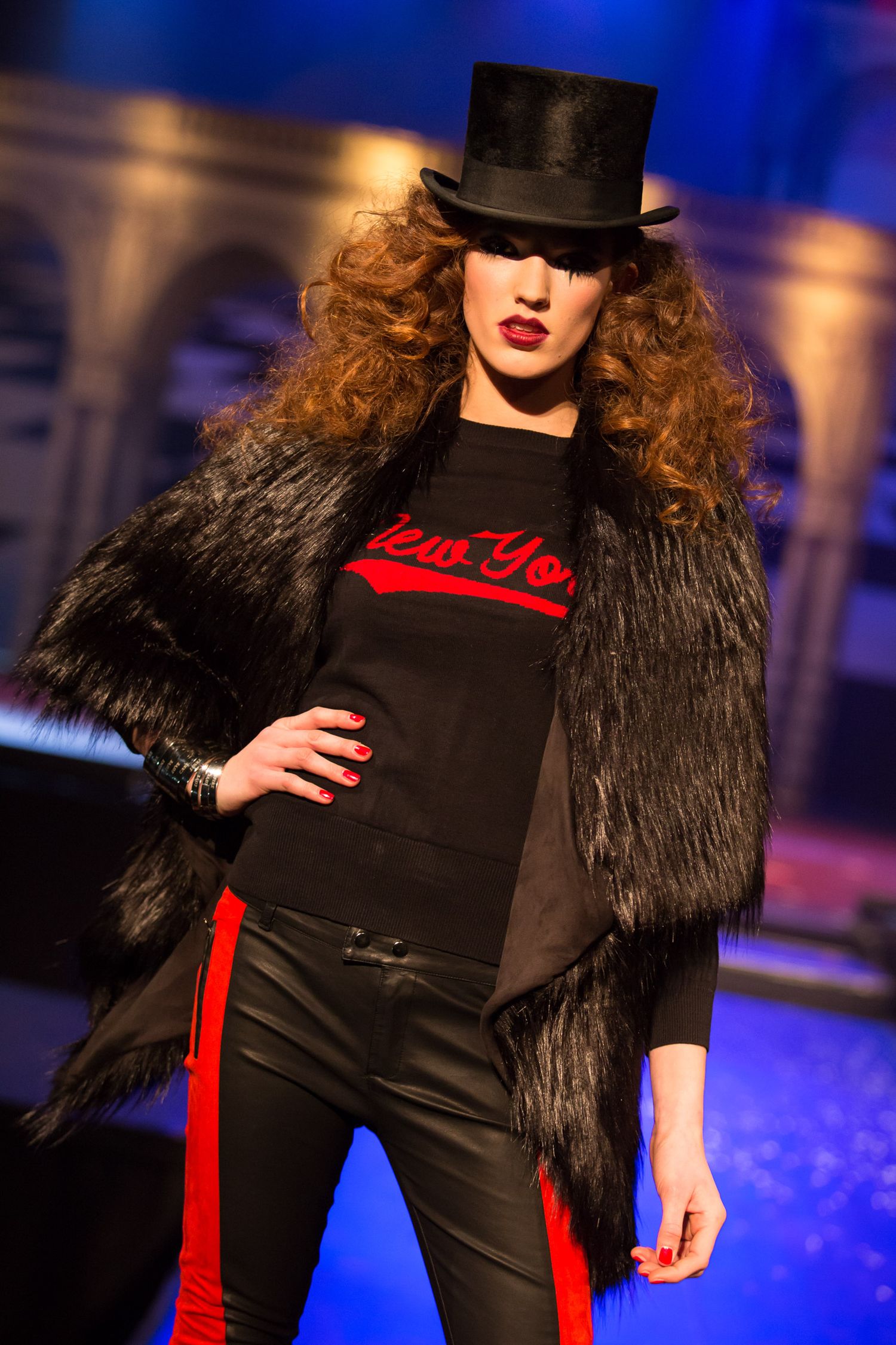 Model wearing black fur coat, top hat, and New York graphic tee with red-striped leather pants on fashion runway.