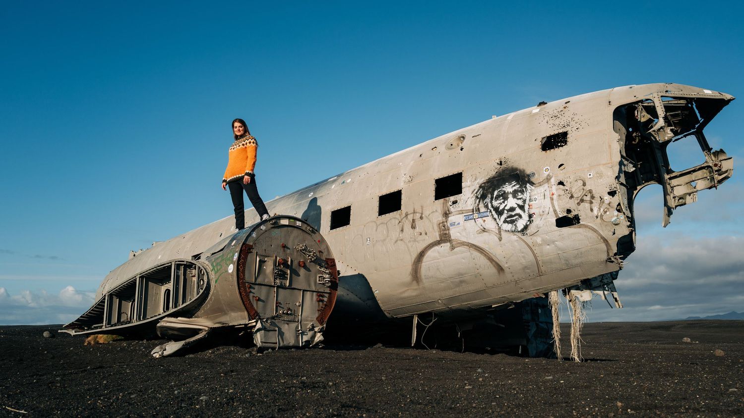 A person stands on top of a crashed DC-3 airplane wreckage against a blue sky on a black sand beach in Iceland.