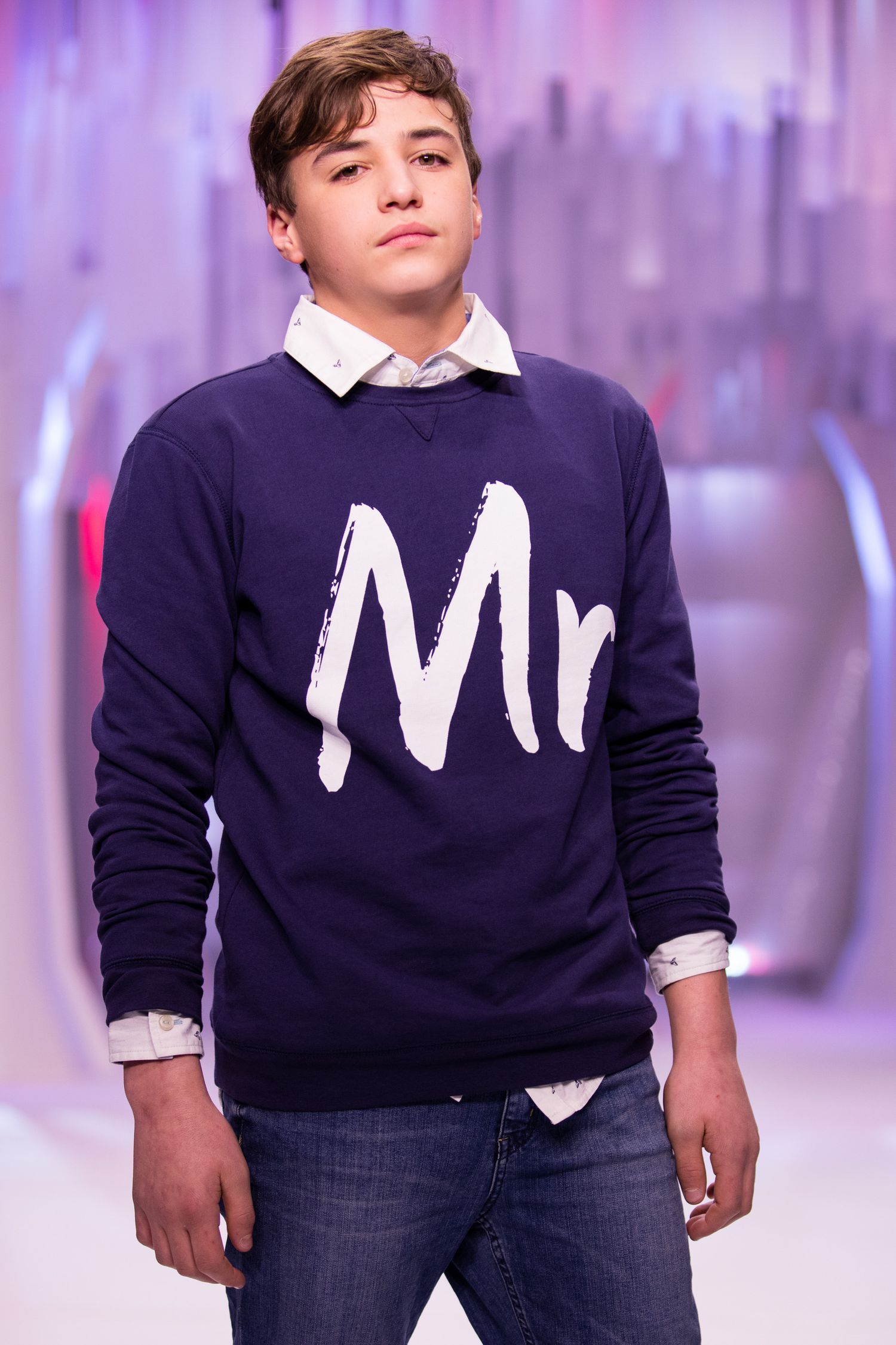 Boy model wearing a navy blue Mr sweatshirt over a white collared shirt with jeans on a fashion runway.