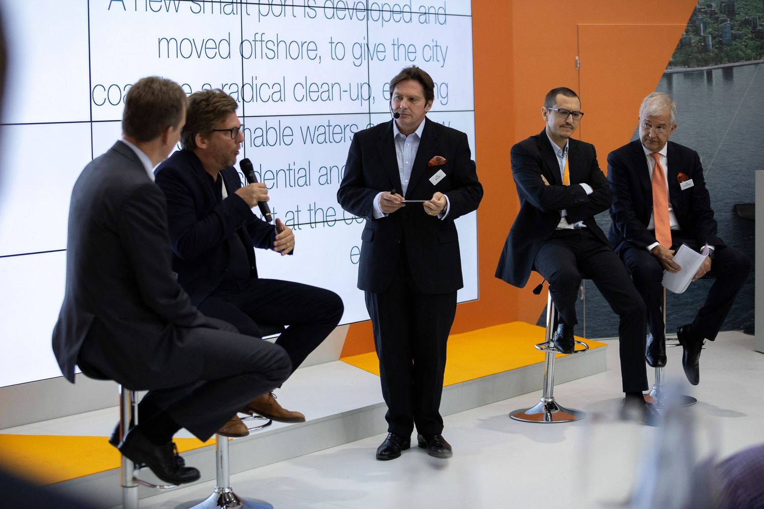 Business professionals in dark suits sit and wait in a modern office hallway with orange and white walls.
