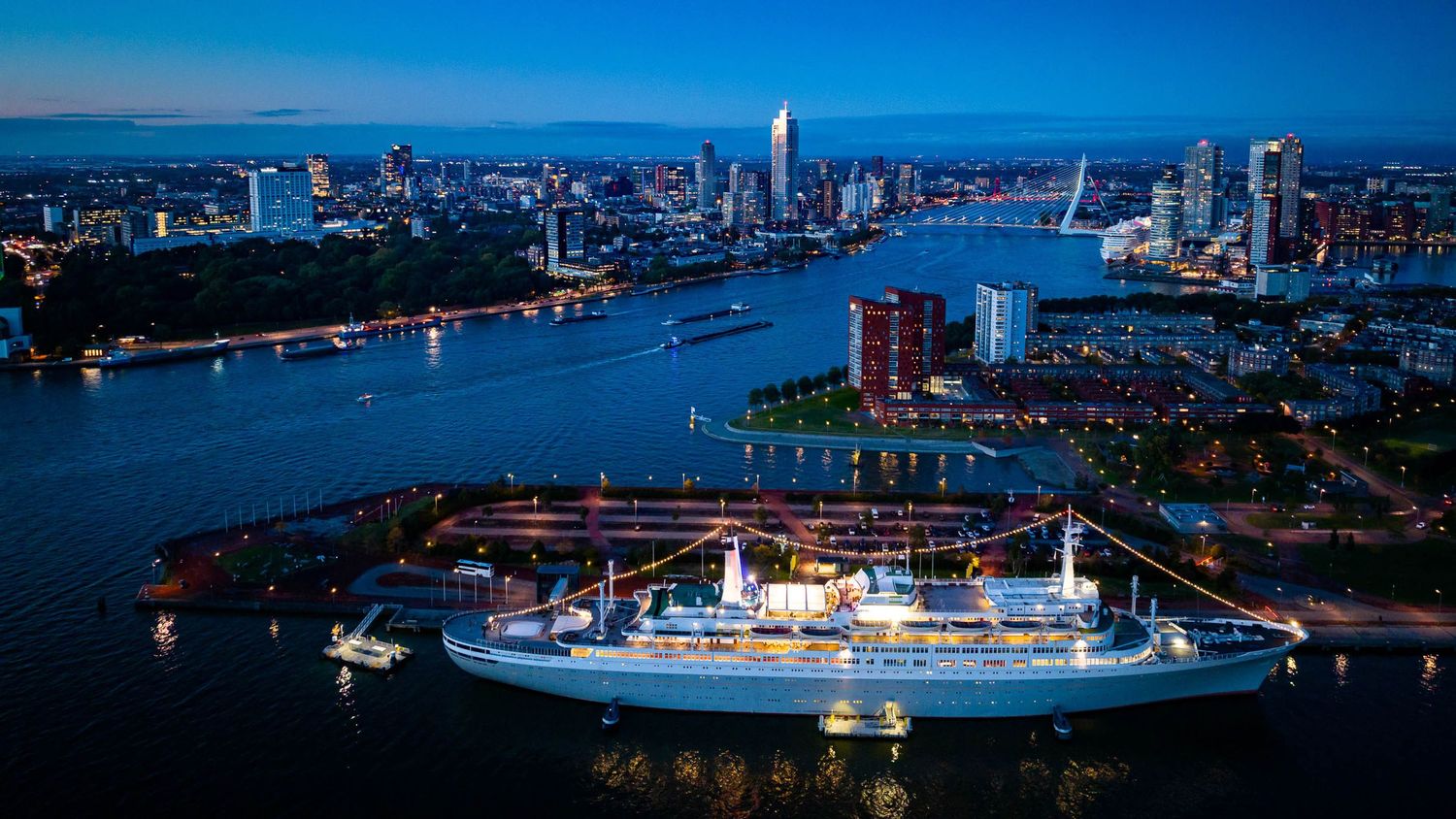 Aerial night view of a cruise ship docked in Rotterdam harbor with illuminated city skyline.