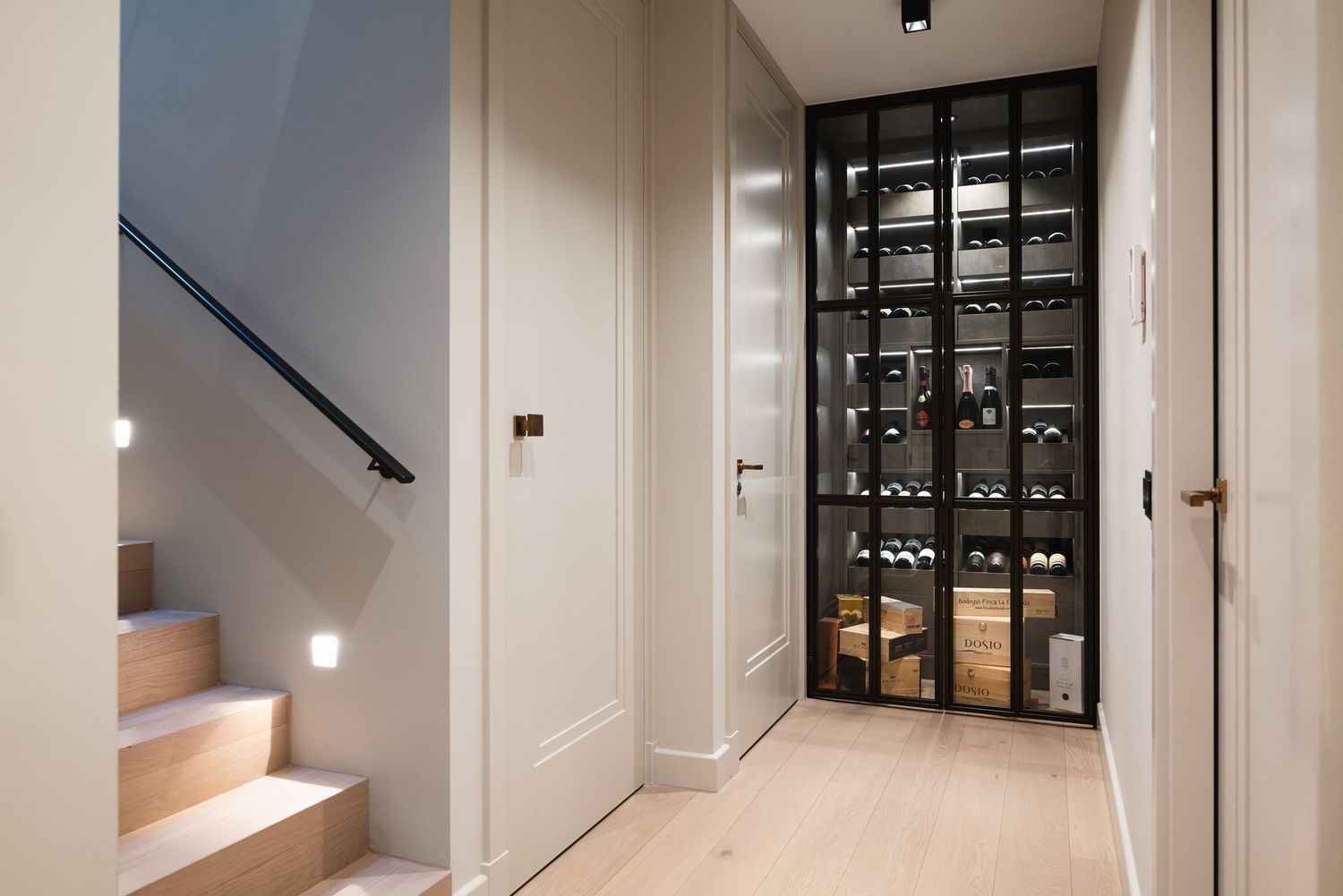 Modern hallway with illuminated staircase and glass-enclosed wine storage display featuring wooden shelving and lighting.
