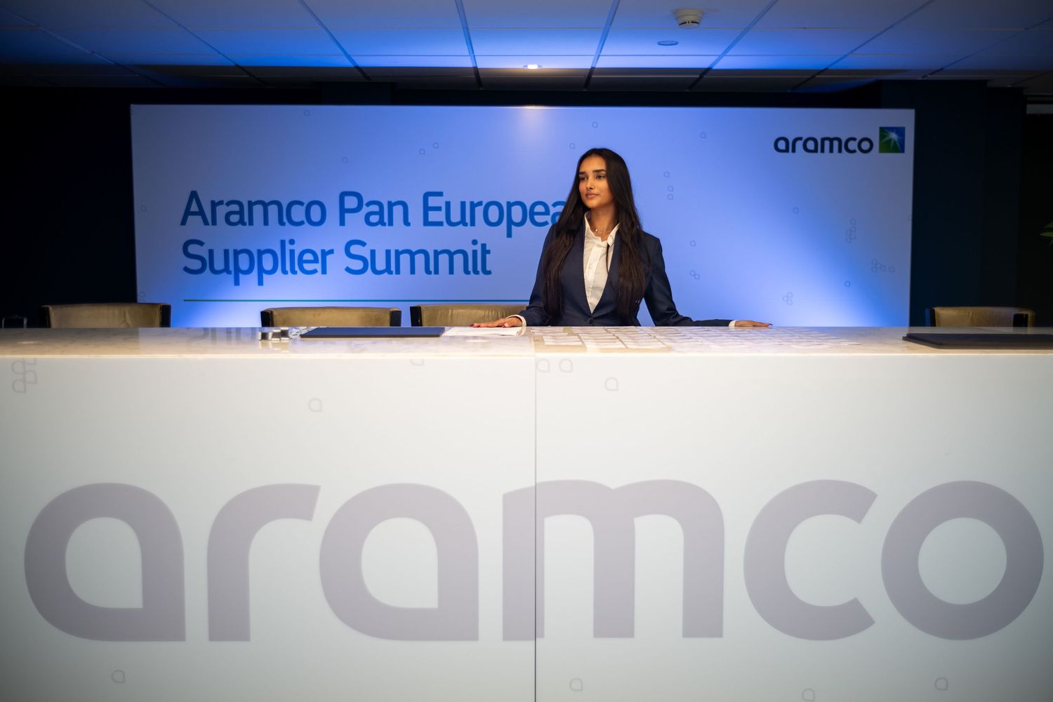 Corporate conference stage with blue lighting and Aramco branding displays.
