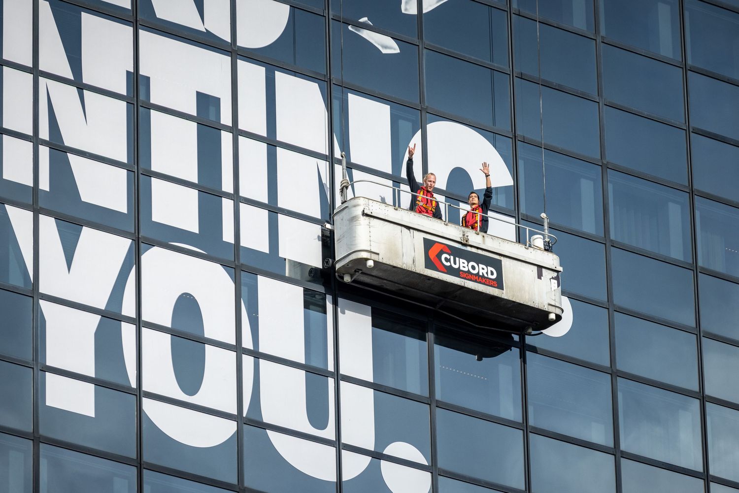 Workers on a suspended scaffold platform clean glass windows of a large building with bold typography signage.