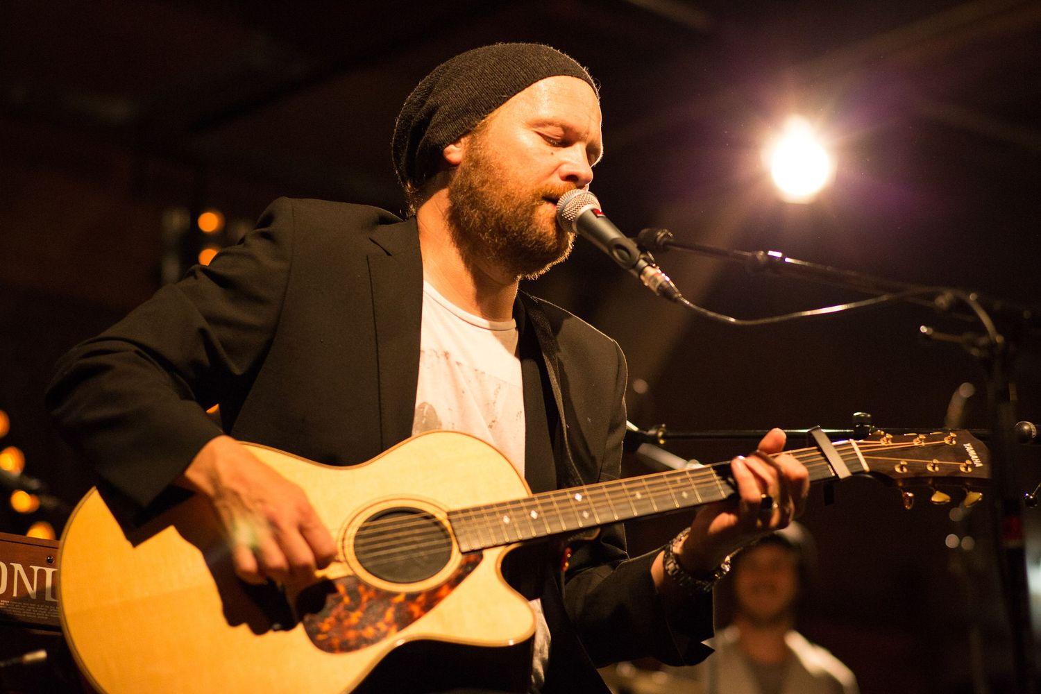 Bearded male musician wearing a black beanie performs live, playing acoustic guitar and singing into a microphone on stage.