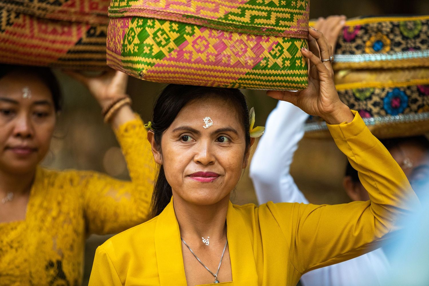 Traditional Balinese woman in yellow dress carrying ornate ceremonial offering basket on her head.