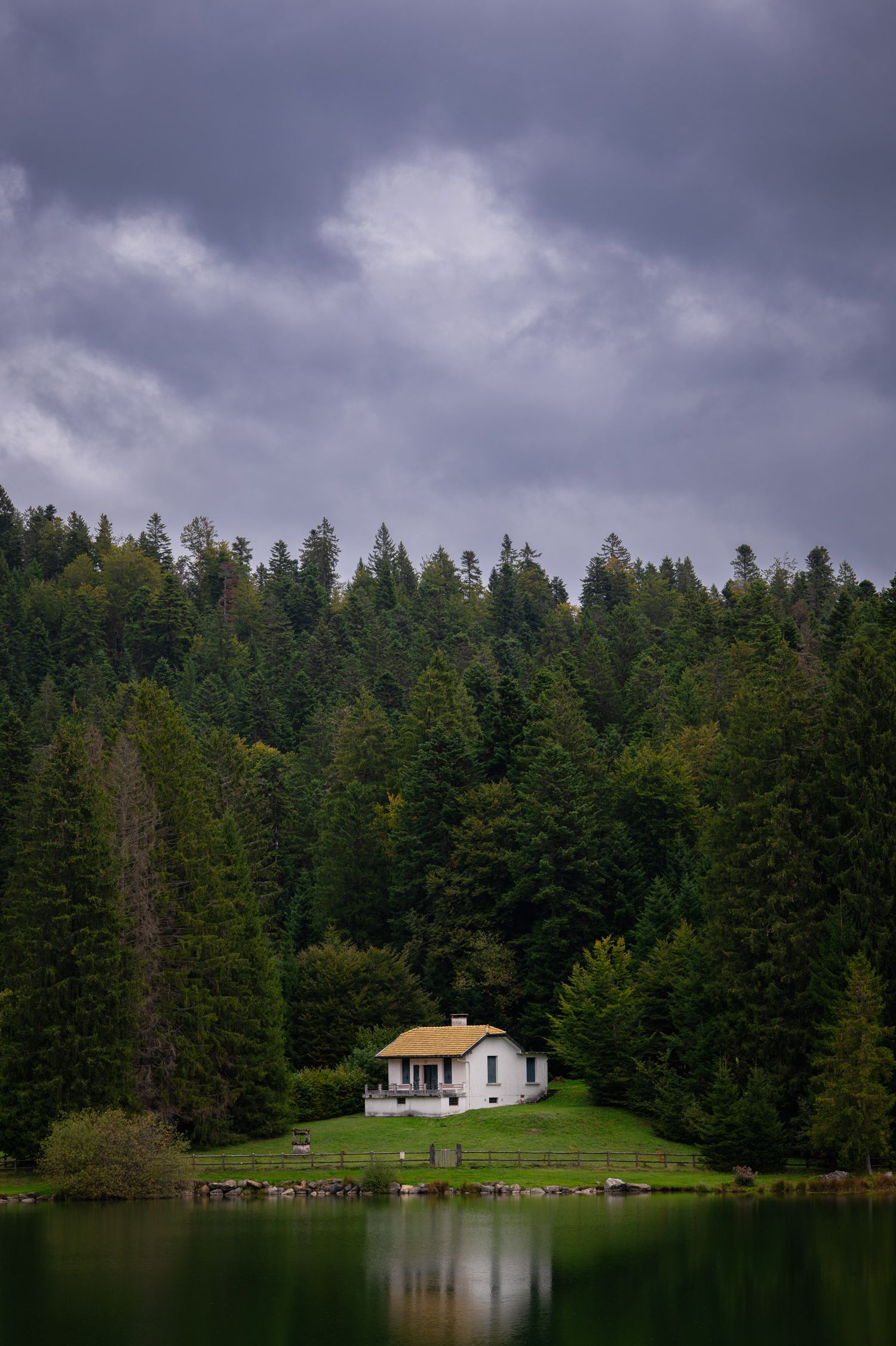 Lone white cottage nestled among pine trees beside tranquil lake under stormy sky.