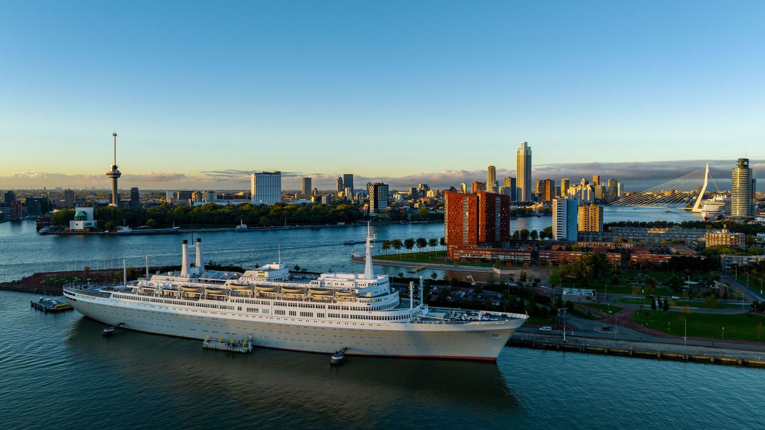 Classic white cruise ship docked in Rotterdam harbor with city skyline and Euromast tower at golden hour.