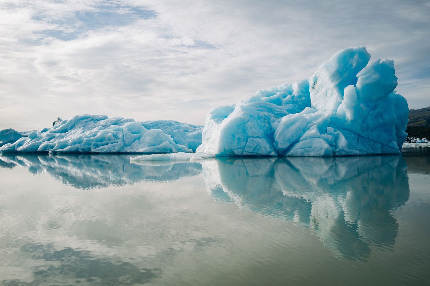 A series of blue glacial icebergs floating in calm water under cloudy skies creates a stunning arctic landscape.