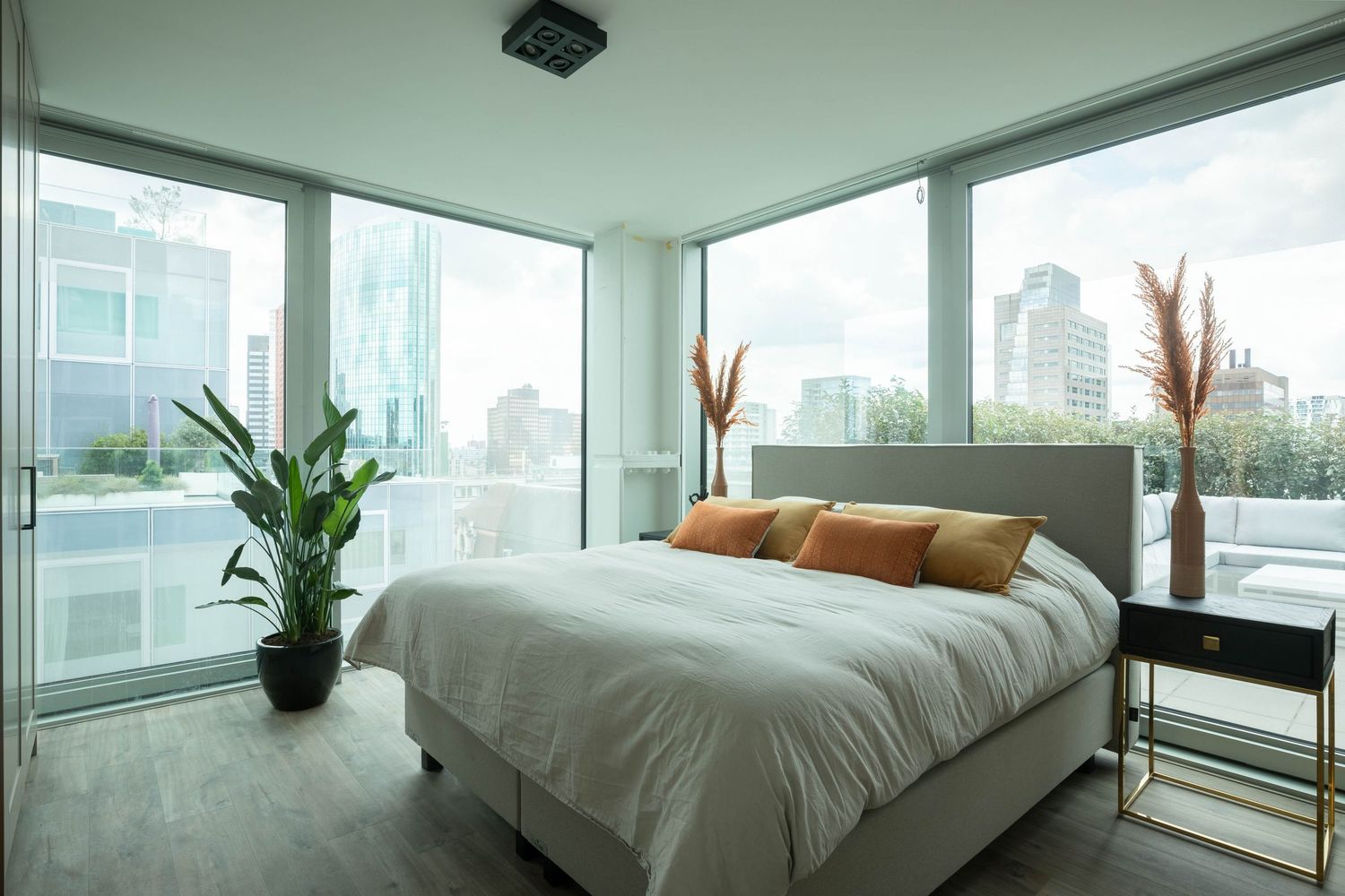 Modern bedroom with panoramic city views features floor-to-ceiling windows, white bedding, and potted plants.