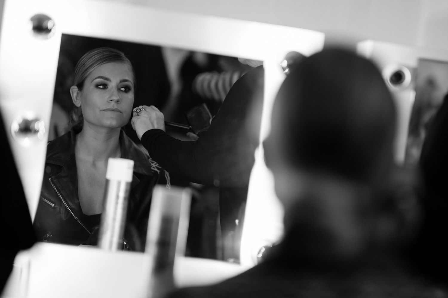 Black and white photo of a woman in a leather jacket getting her hair done, reflected in a vanity mirror.