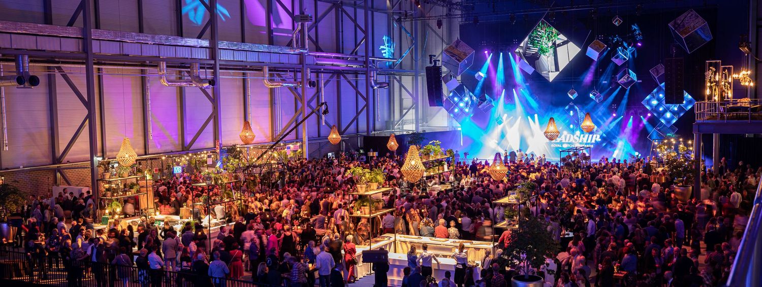 A large crowd gathers at an outdoor music festival with colorful stage lighting and concert atmosphere at night.