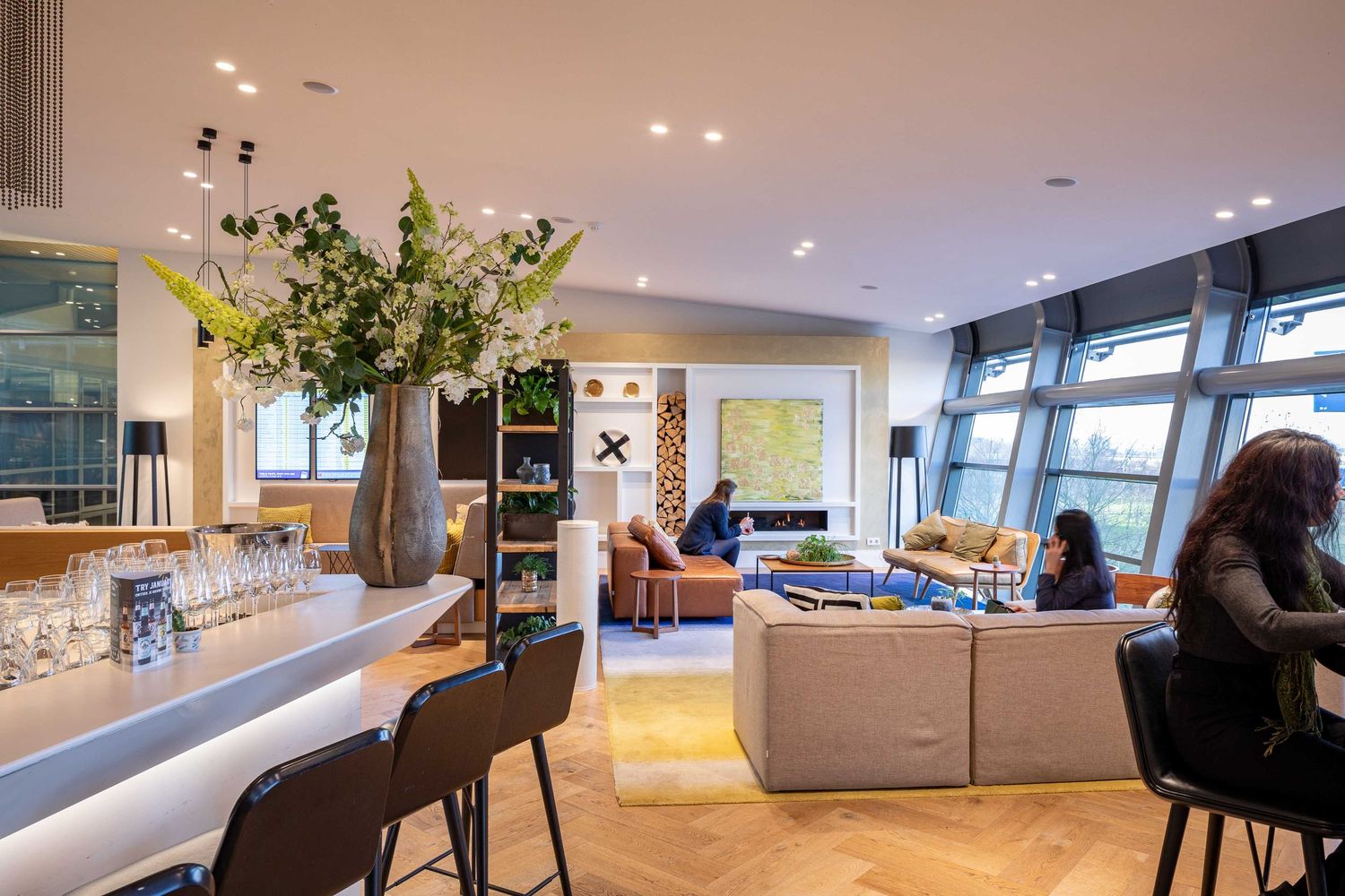 Modern lounge area with bar seating, comfortable couches, and tall floral arrangement in an open-concept space.