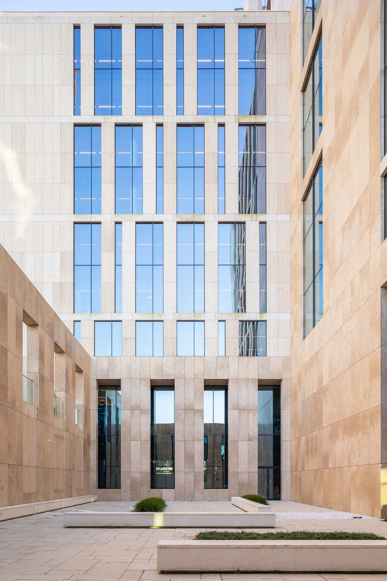 Modern office building exterior with beige stone walls and tall glass windows reflecting blue sky.