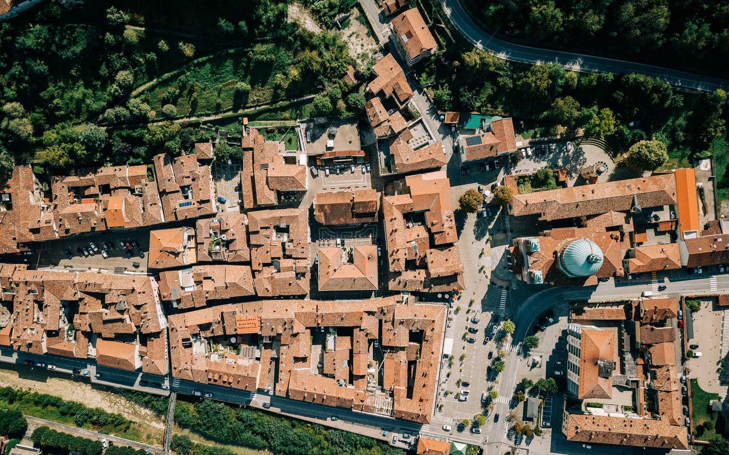 Aerial view of a dense Italian medieval town with terracotta rooftops surrounded by lush green trees.