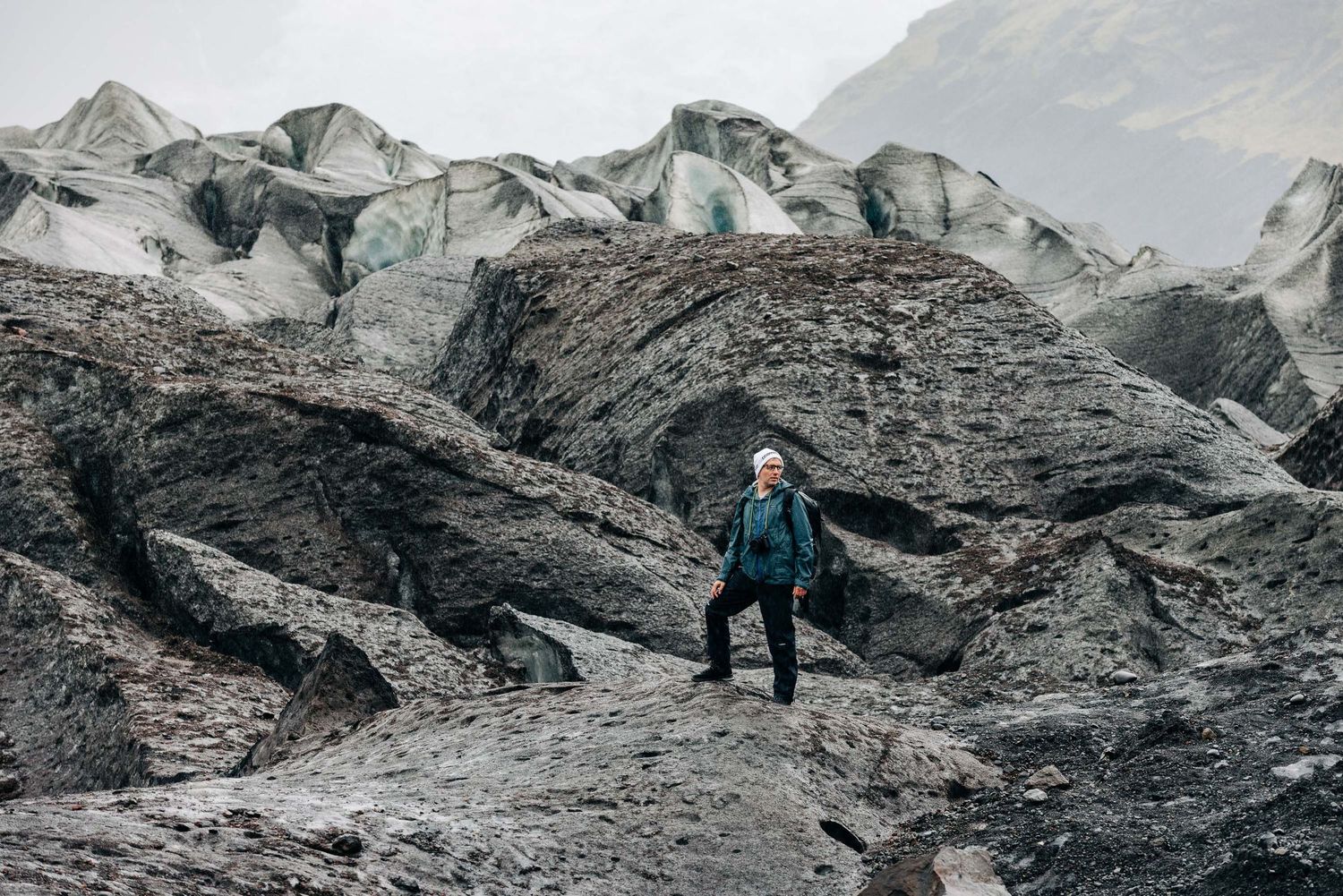 A hiker traverses a rugged mountain trail with dramatic snow-capped peaks and glaciers in the background.