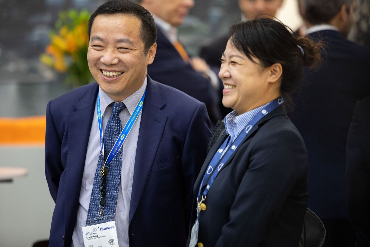 Two conference attendees wearing blue lanyards and business attire share a cheerful moment at a professional event.