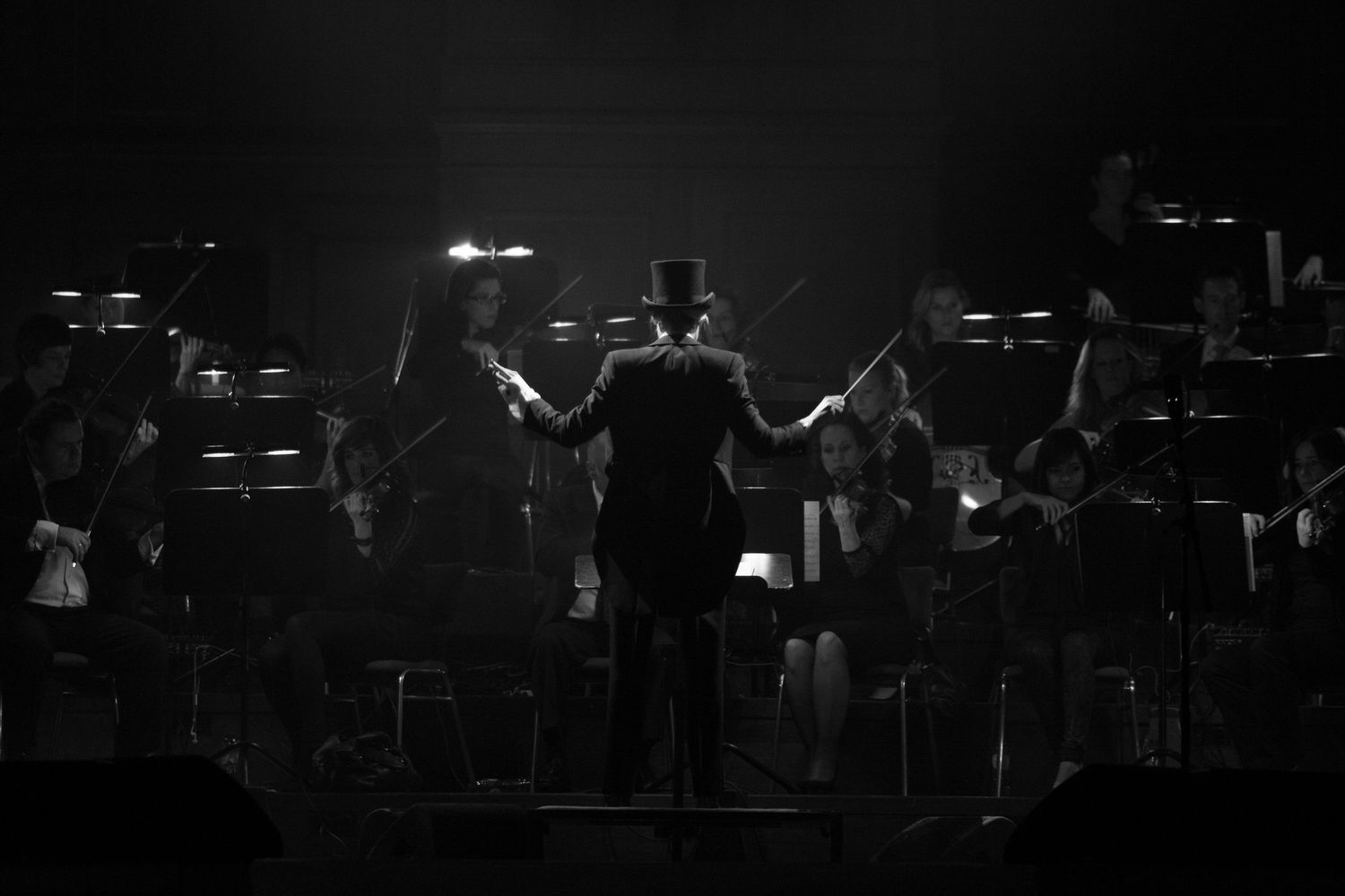 Black and white photo of a conductor in top hat leading an orchestra on a dimly lit stage.