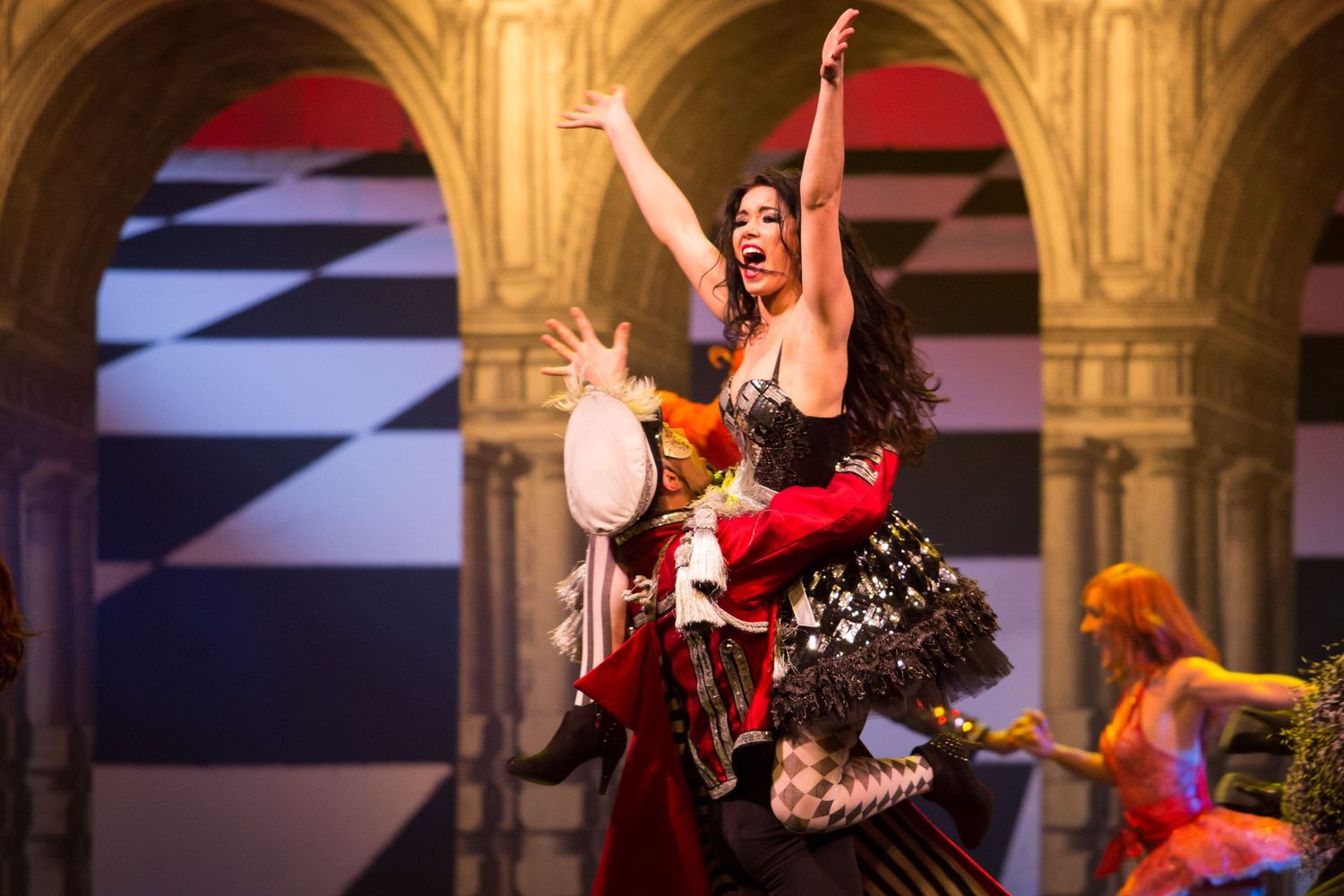 A dramatic performer in an elaborate costume dances on stage during a theatrical production with a checkered backdrop.