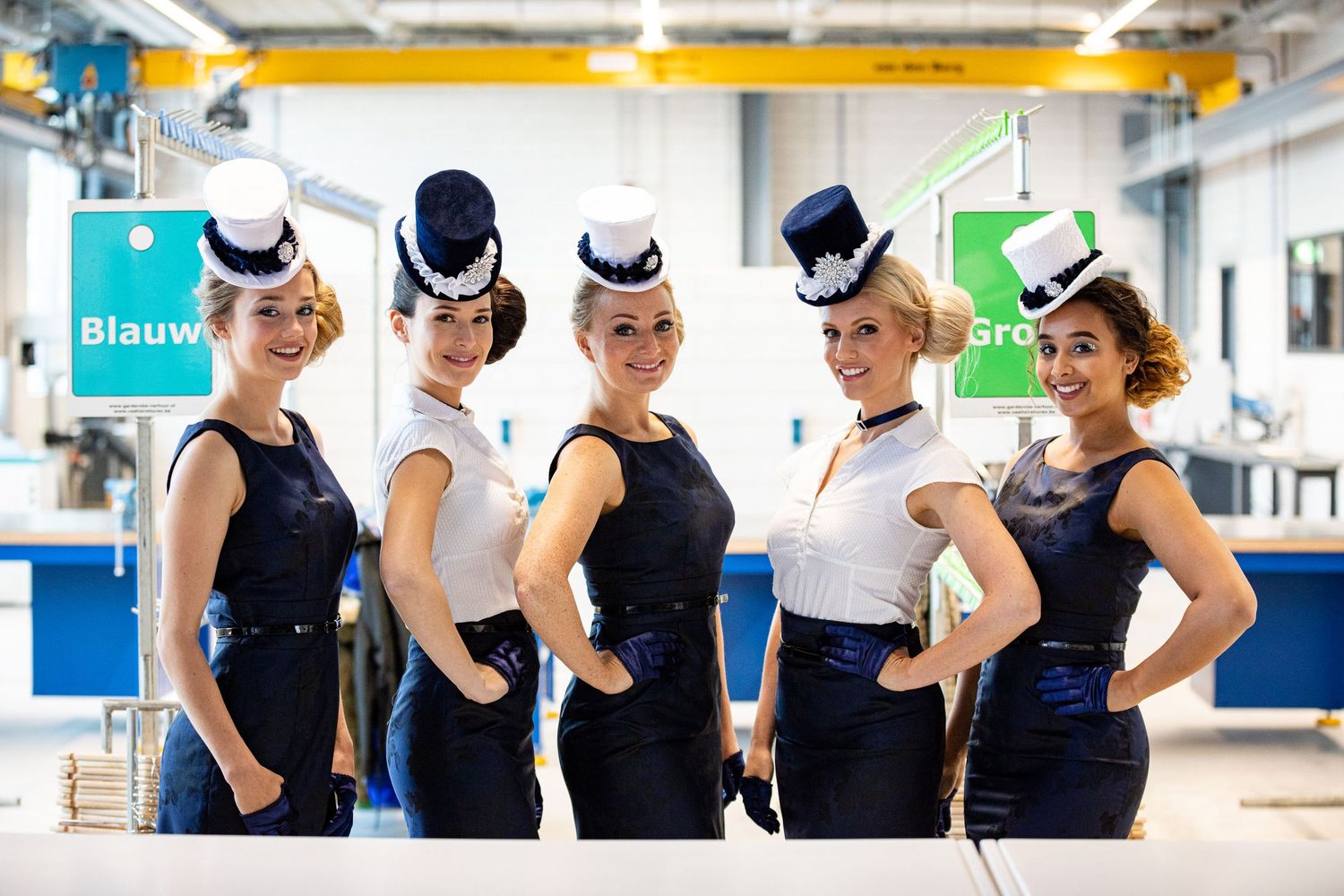 A group of dancers in matching black tops and white hats perform a choreographed dance routine in a studio space.