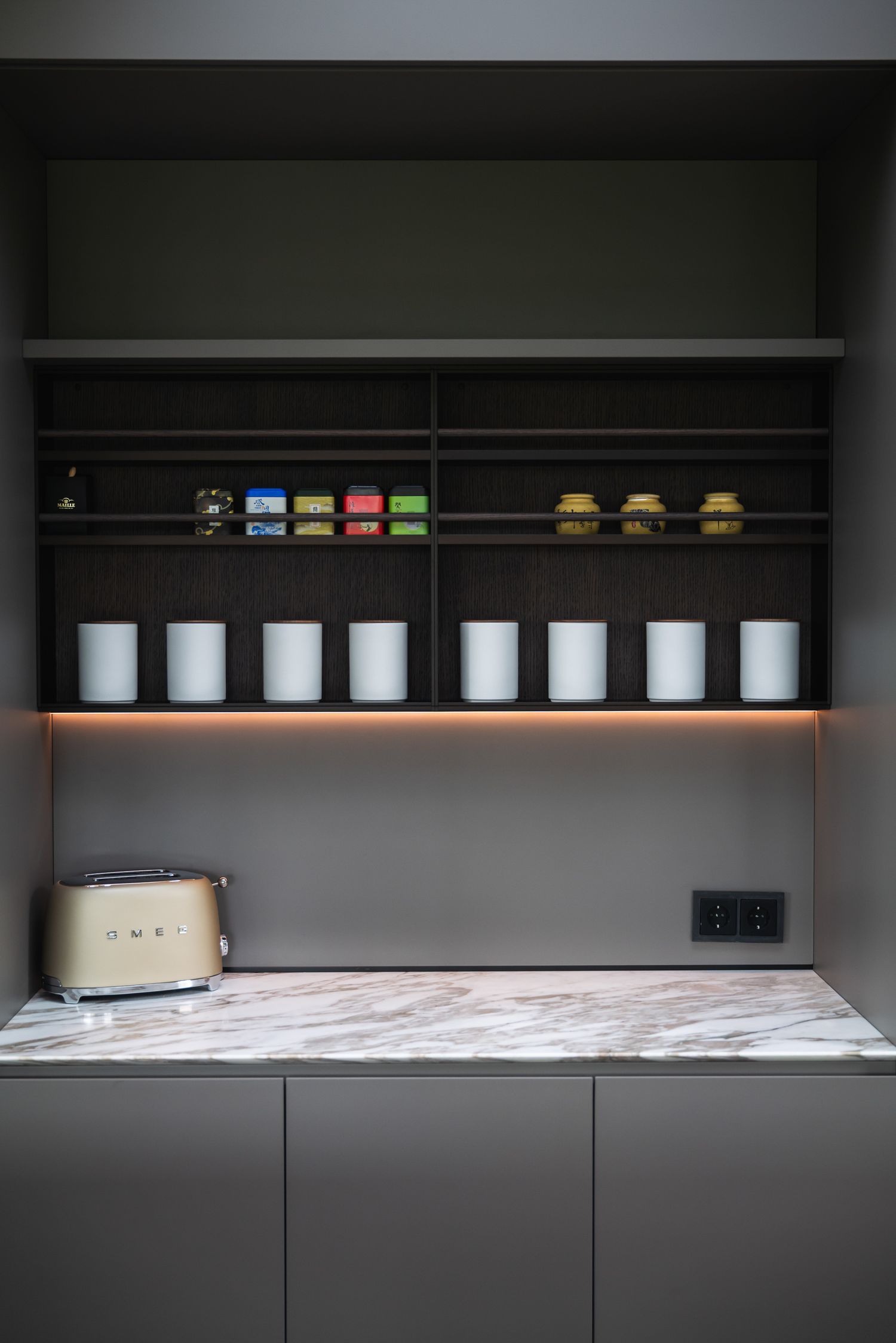 Minimalist kitchen shelving with dark cabinets displays white ceramic containers and colorful tea boxes on wooden ledges.