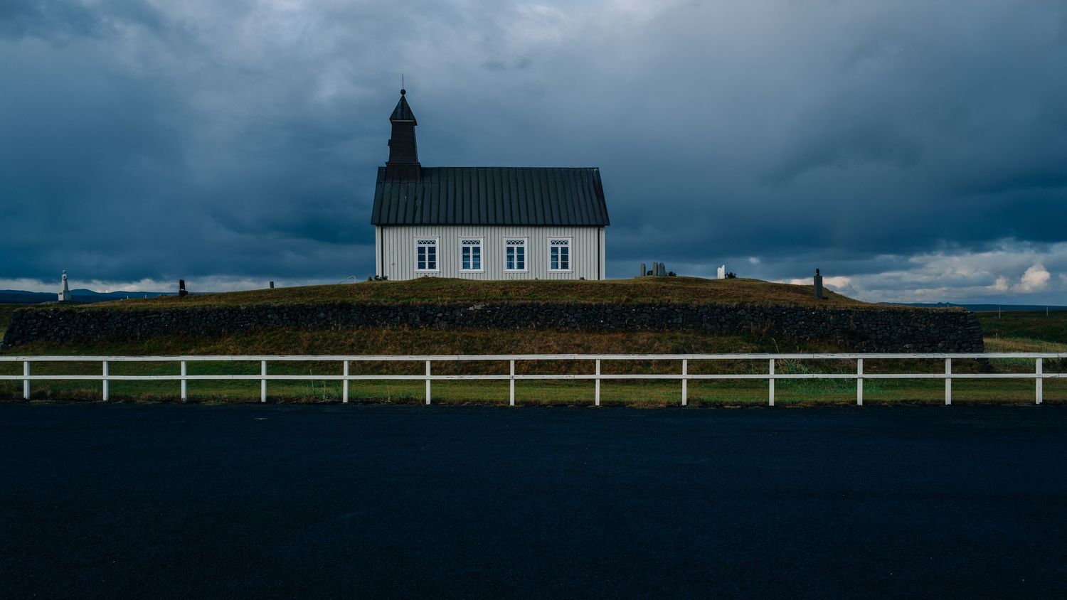 A small white church sits on a hilltop against a dramatic stormy sky with dark clouds looming overhead.