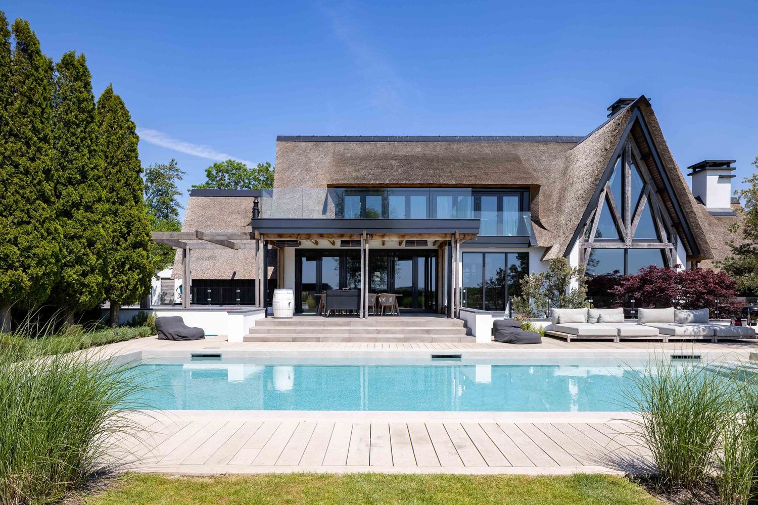 Modern luxury villa with thatched roof design featuring large windows and swimming pool surrounded by wooden deck.