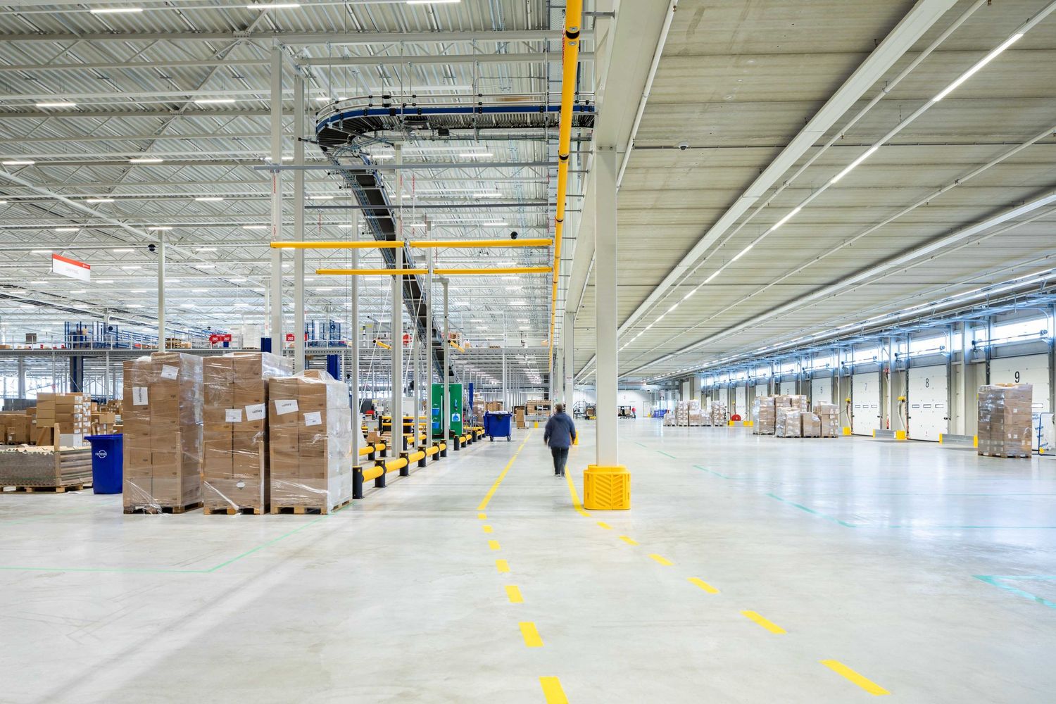 Large empty warehouse interior with white walls, fluorescent lighting, and yellow safety markings on the concrete floor.