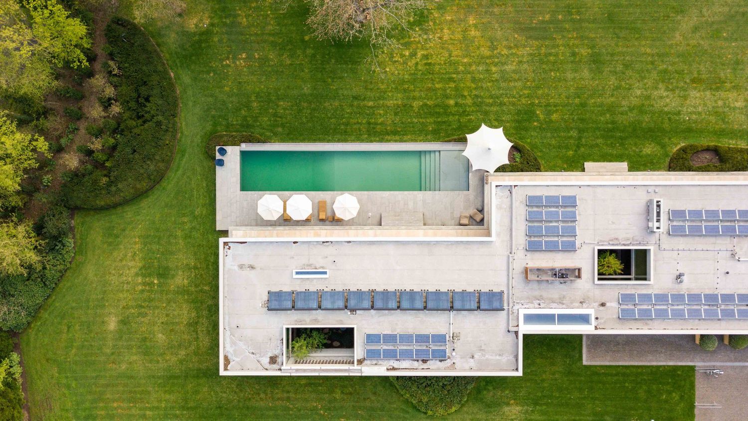 Aerial drone view of a modern swimming pool and tennis courts surrounded by lush green landscaping.