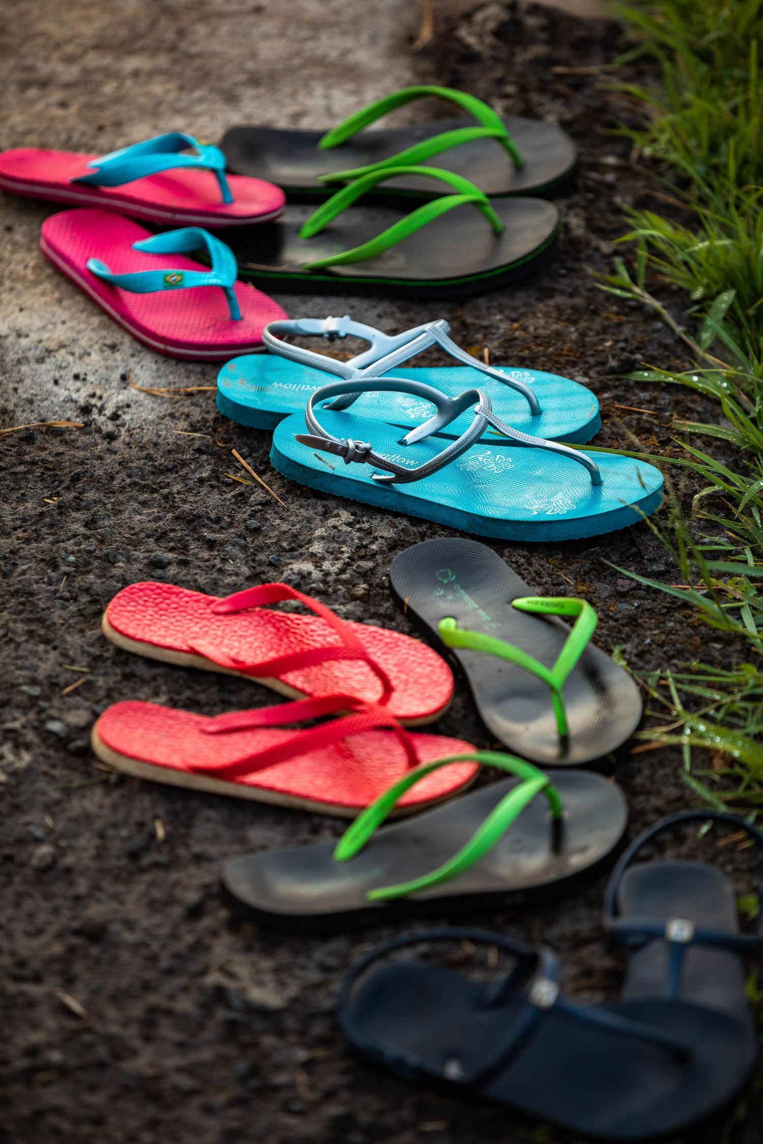 Colorful flip flops arranged in pairs on dark ground with grass in background.