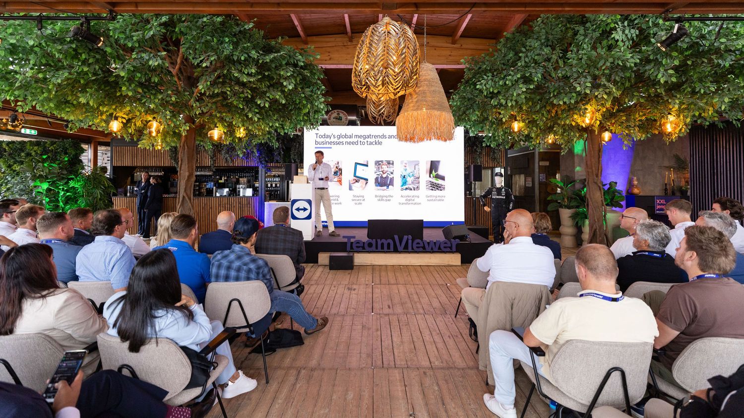 Wide shot of outdoor business presentation with audience seated under wooden pergola.