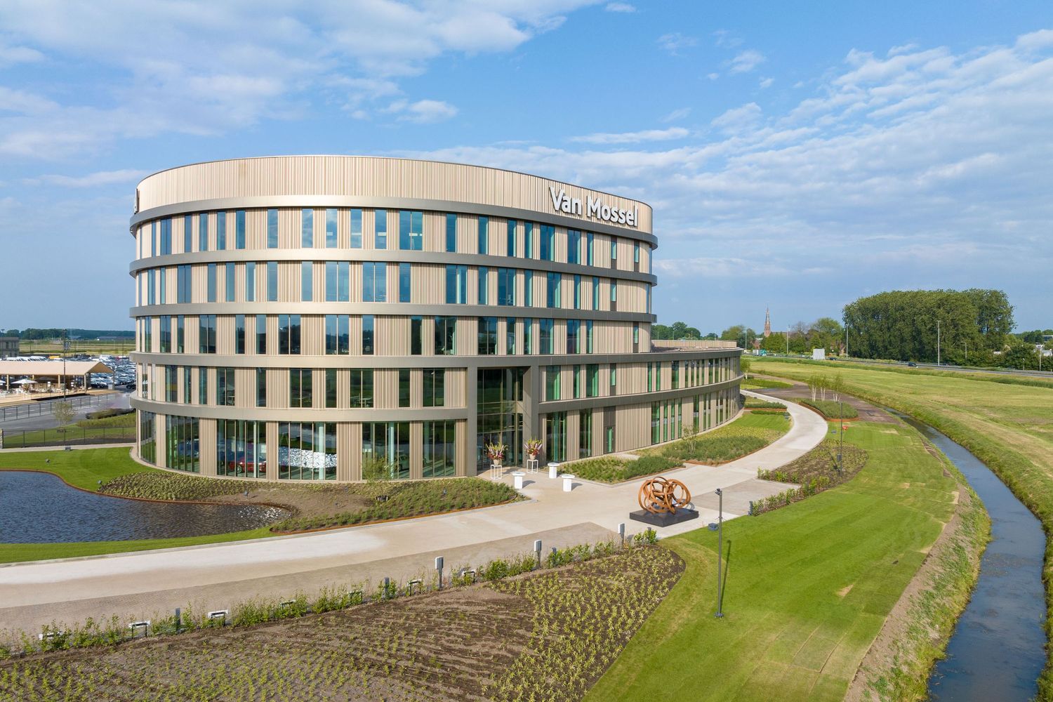 Modern curved office building with glass facade sits along a winding path near green landscaping and water features.
