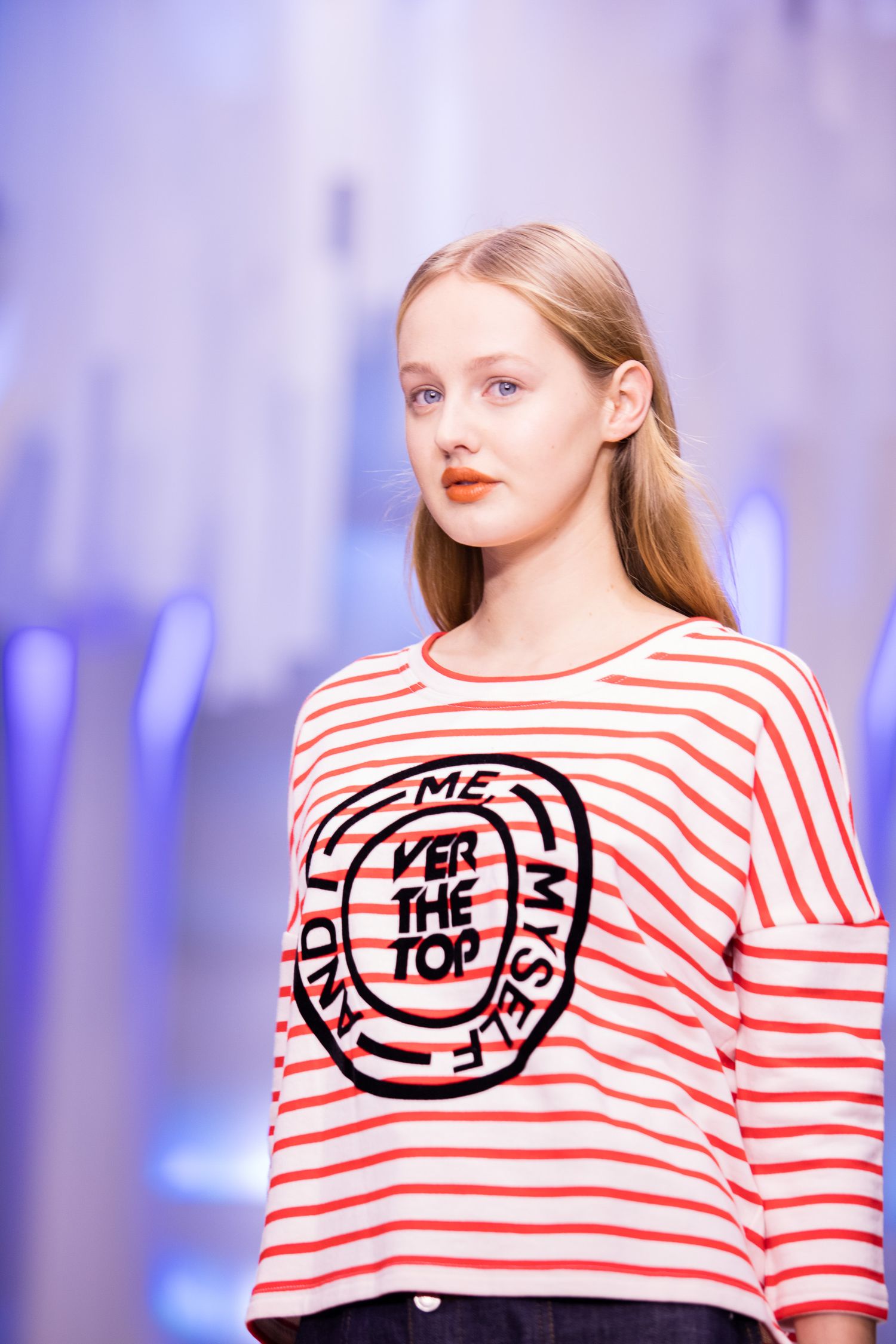 Female model on runway wearing red and white striped top with 'Over The Top' graphic print at fashion show.