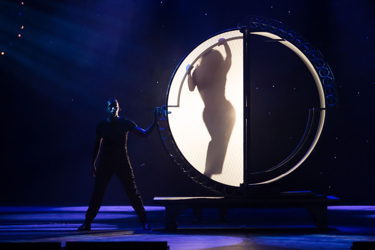 Silhouette of a performer in a glowing circular frame on a dark stage with dramatic lighting during a live show.