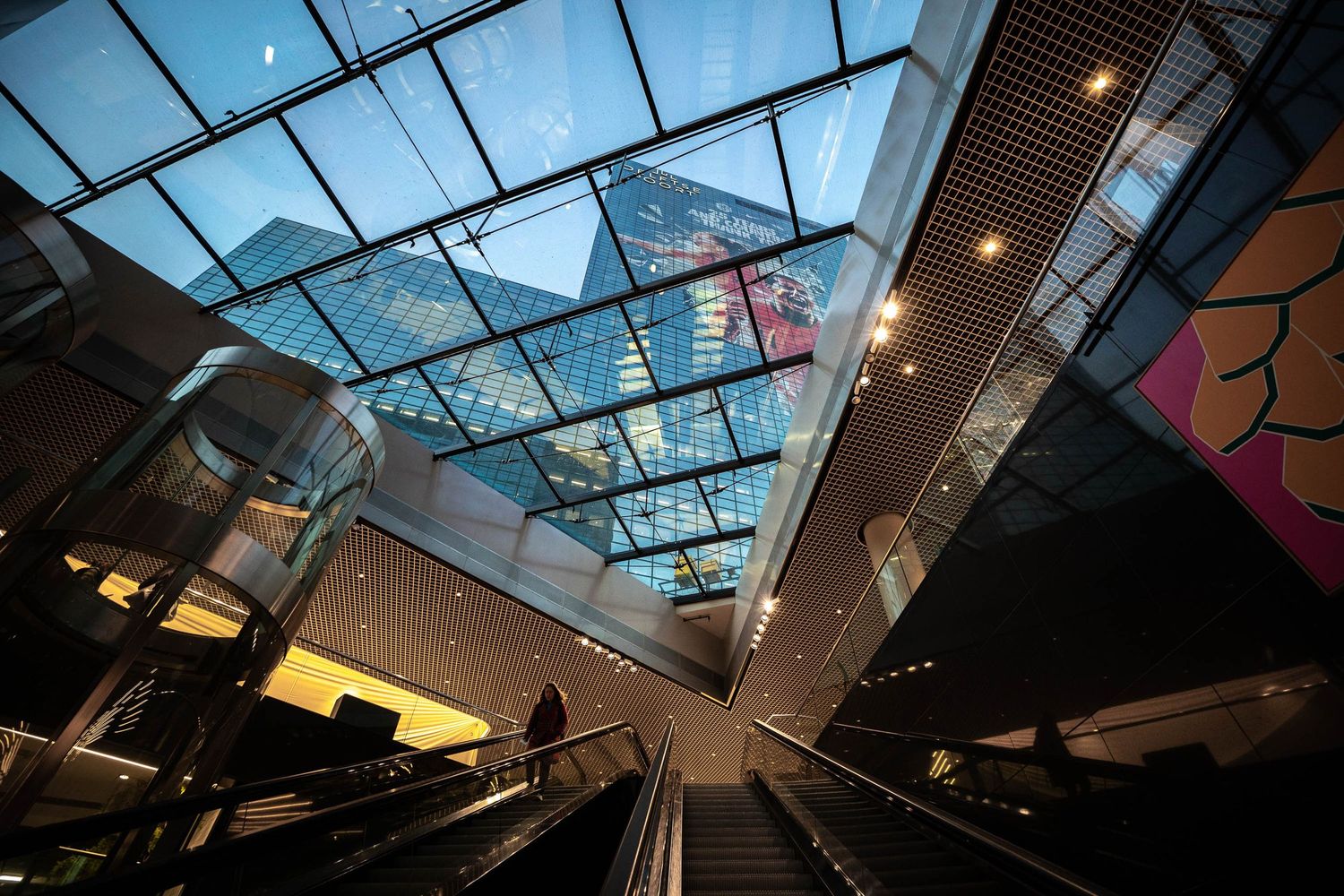 Modern shopping mall interior with glass ceiling, escalators, and contemporary architecture reflecting city buildings above.