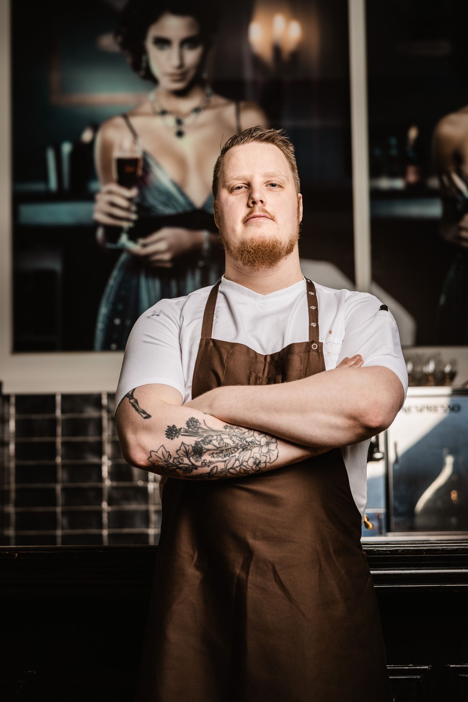 Professional chef in brown apron and white shirt stands confidently in restaurant kitchen with crossed arms and tattoos.