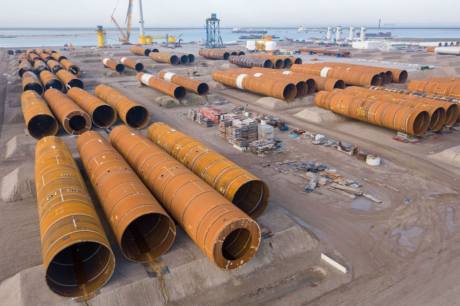 Aerial view of large steel wind turbine monopile sections stored at an industrial port facility.
