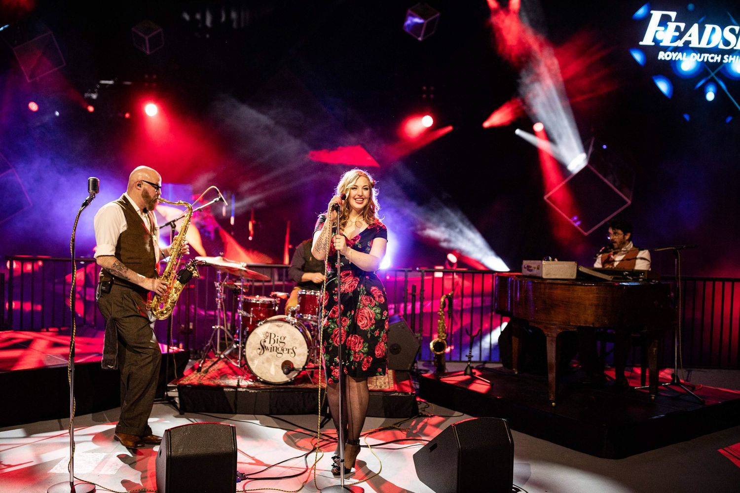 Musicians perform on stage with colorful red lighting and fog effects at a live concert venue.