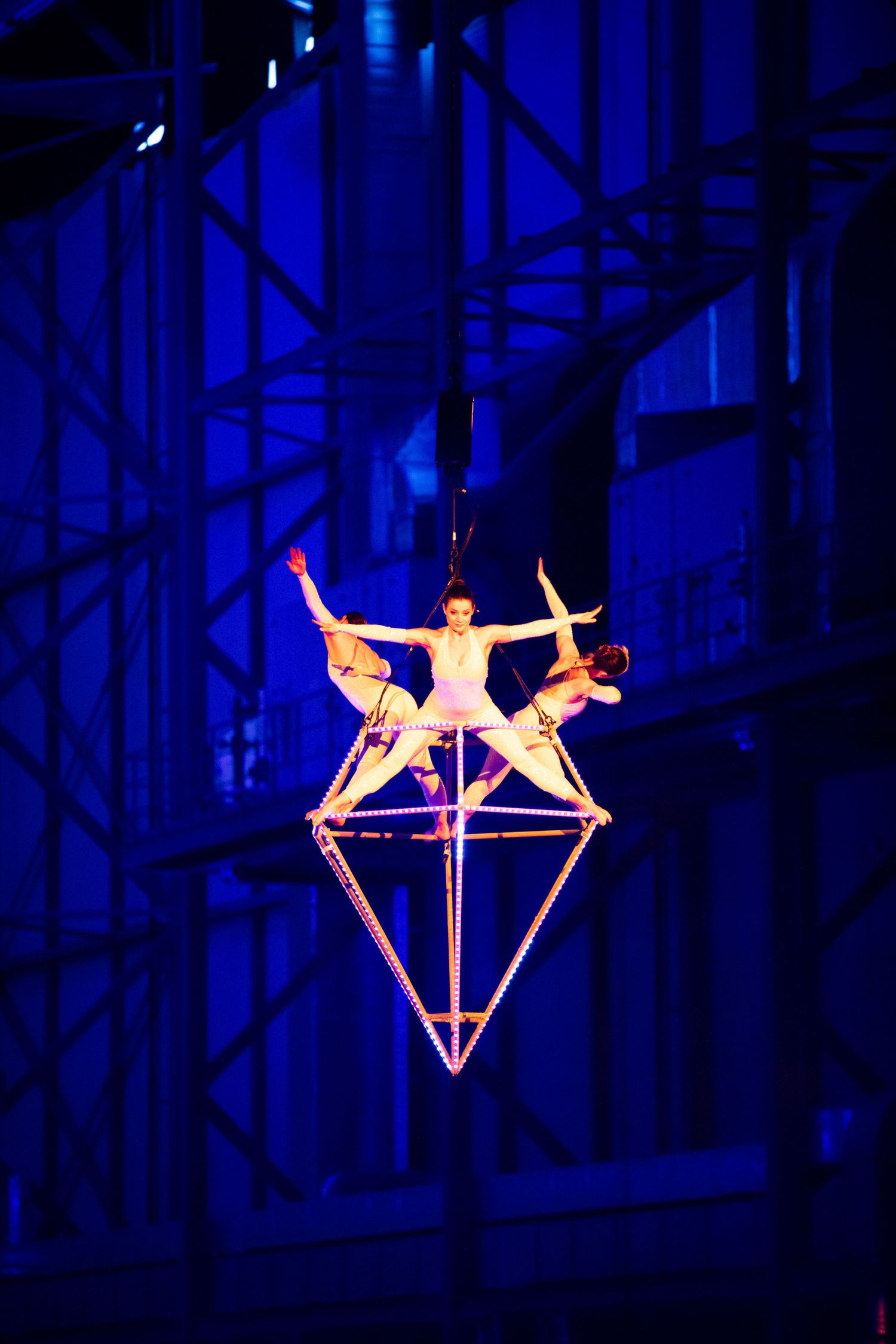 Aerial acrobats perform a dramatic diamond-shaped pose on an illuminated platform against a deep blue stage background.