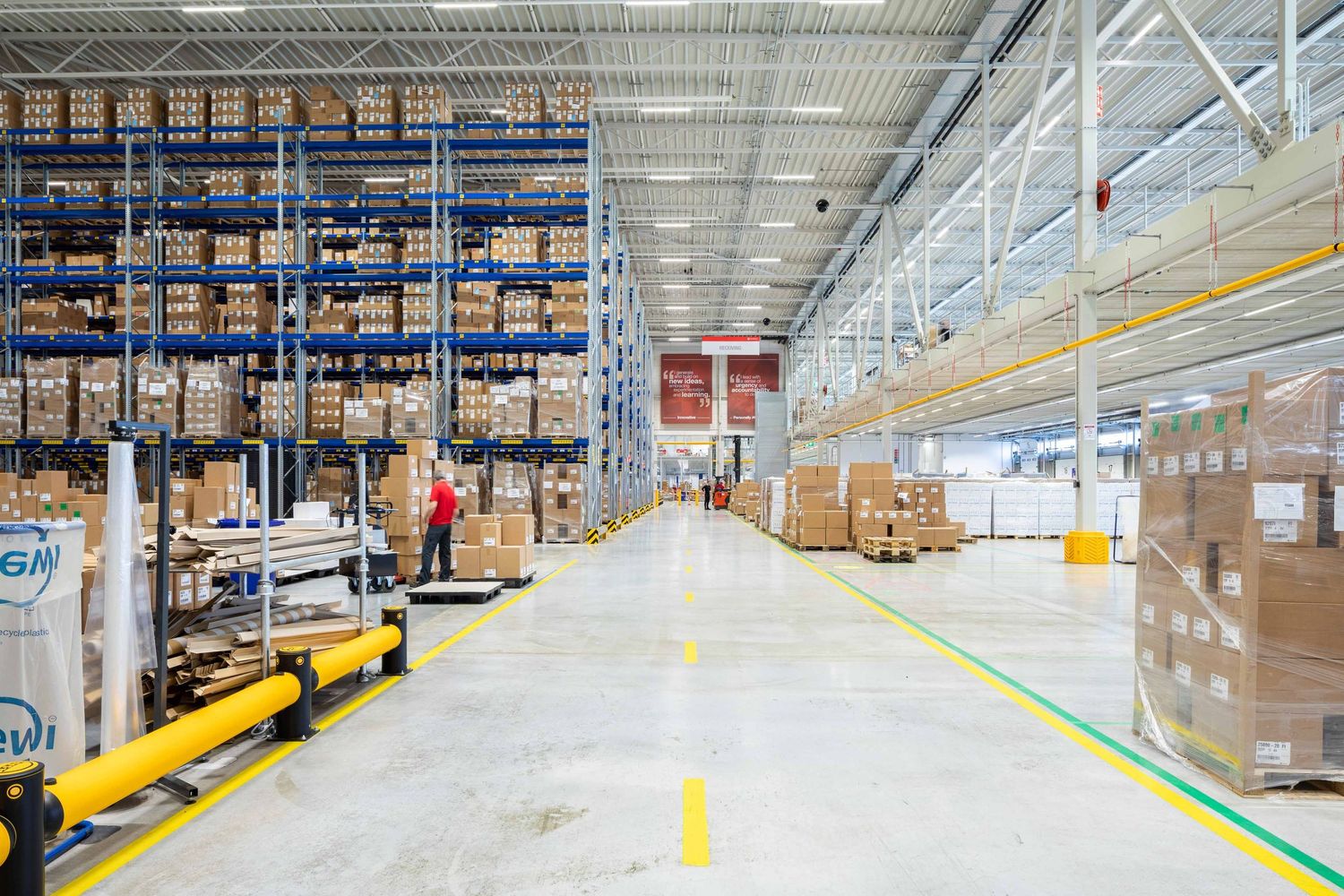 Long rows of tall storage racks filled with boxes line the aisles of a large industrial warehouse facility.