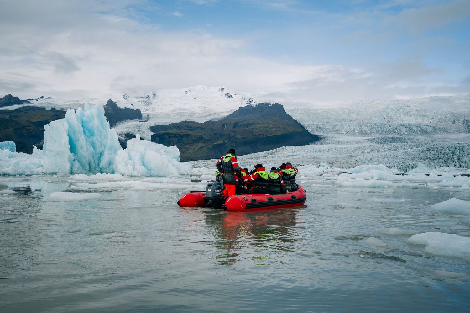 A red inflatable boat navigates through icy waters surrounded by glaciers and floating ice under a cloudy sky.