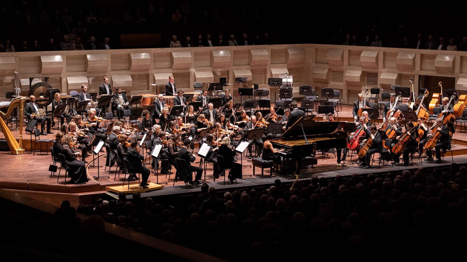 Wide panoramic view of classical orchestra arranged on illuminated concert hall stage during performance.