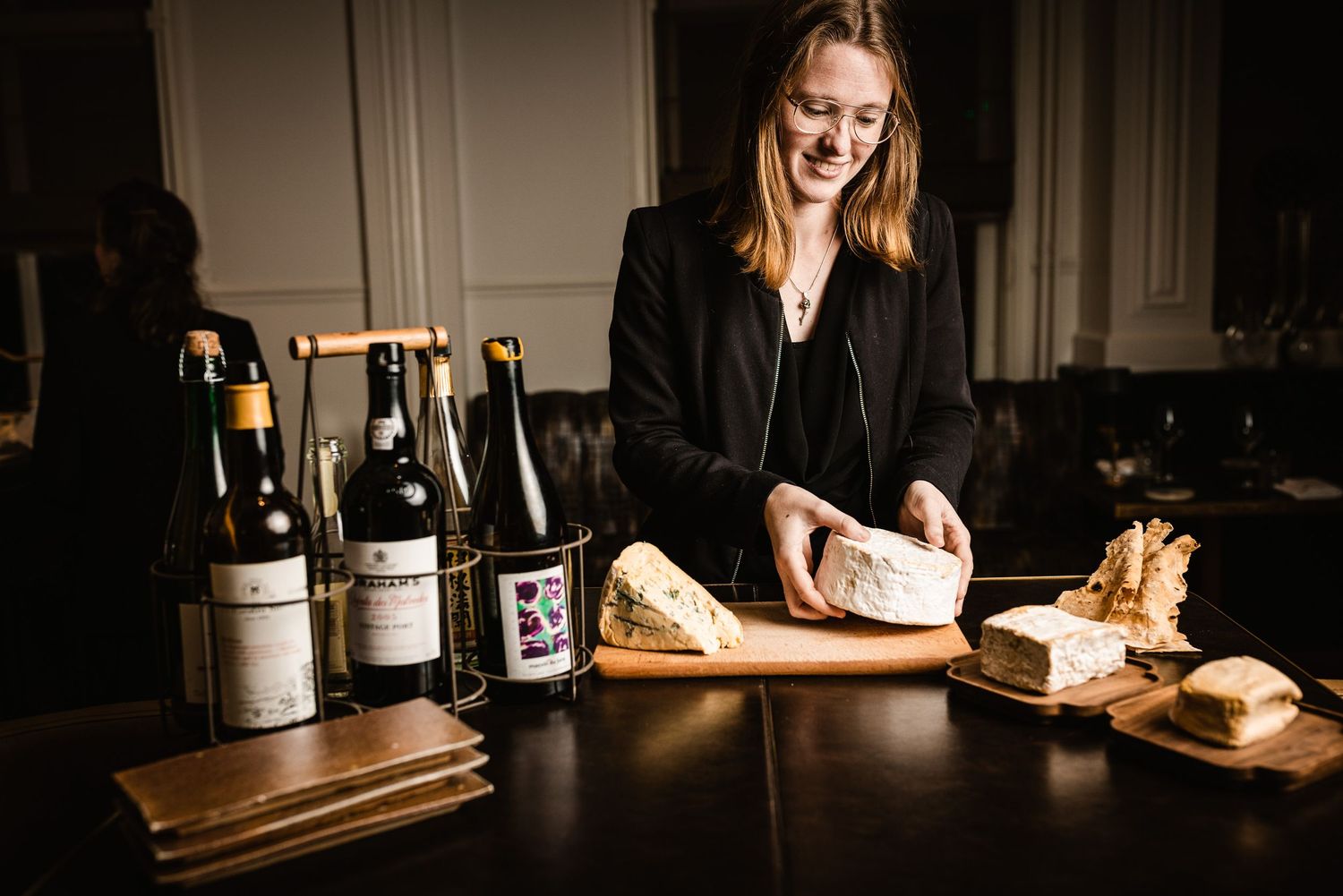 A person in black attire slices bread on a wooden board next to several wine bottles in a dimly lit room.