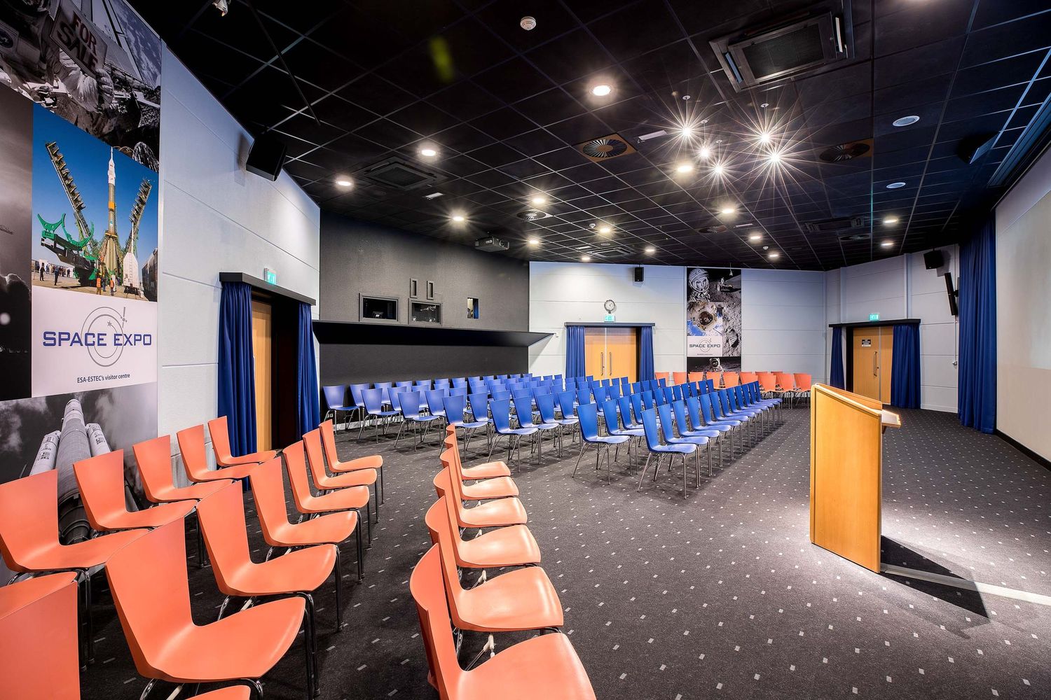 Modern conference room at Space Expo with orange and blue chairs, podium, and space-themed wall banners.